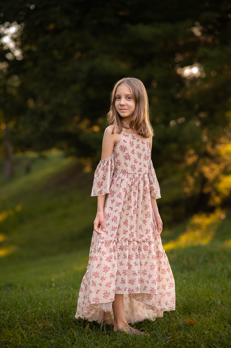 a young girl in an off the shoulder flowered gown stands in lush green grass surrounded by evergreen trees