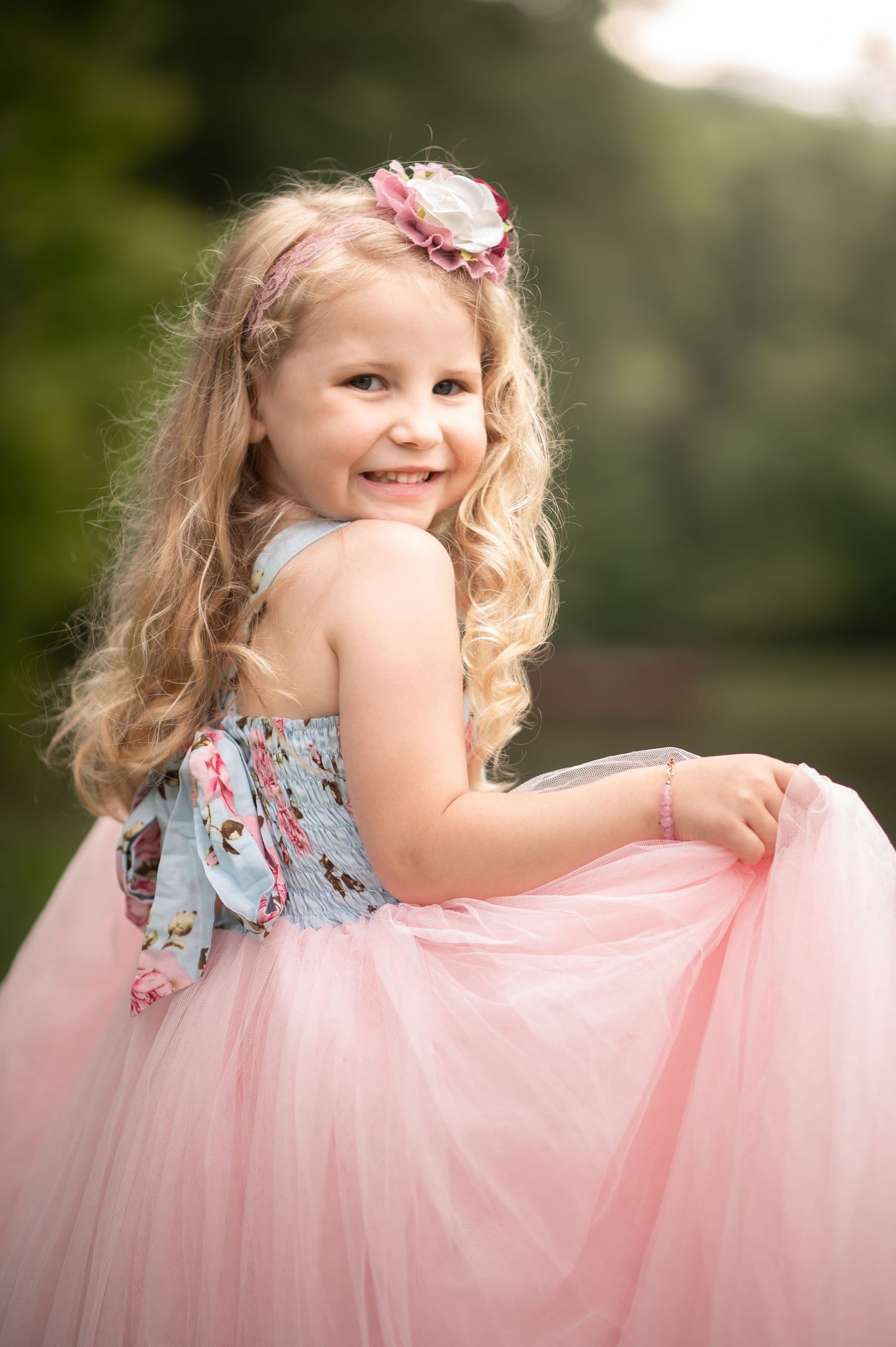 a little girl in a pink and blue floral dress laughs and twirls, with trees in the background