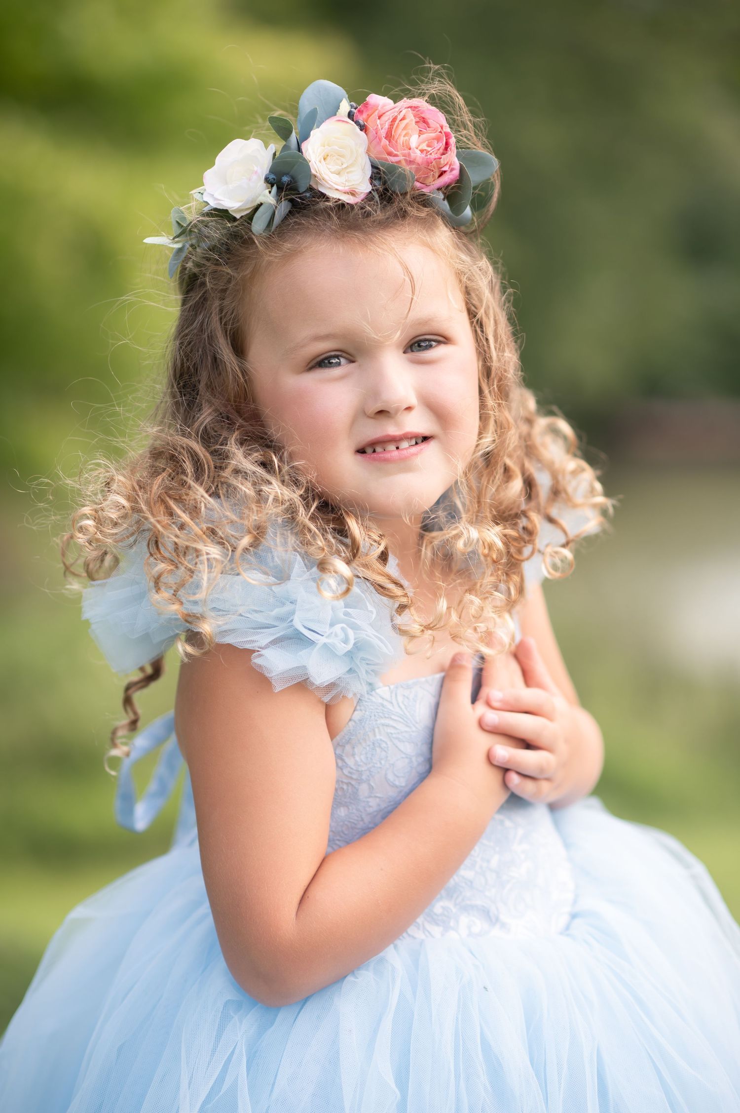 a pretty little girl with curly hair in a blue dress and floral crown smiles for a portrait with green trees in the background
