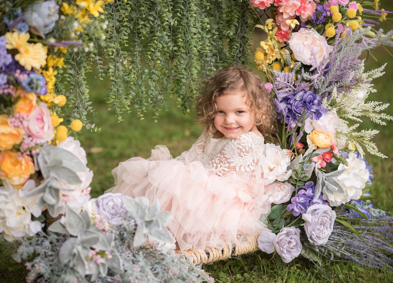 a little girl with curly hair and a fluffy pink and white dress sits in a ring of spring flowers and hanging ivy and smiles for a portrait