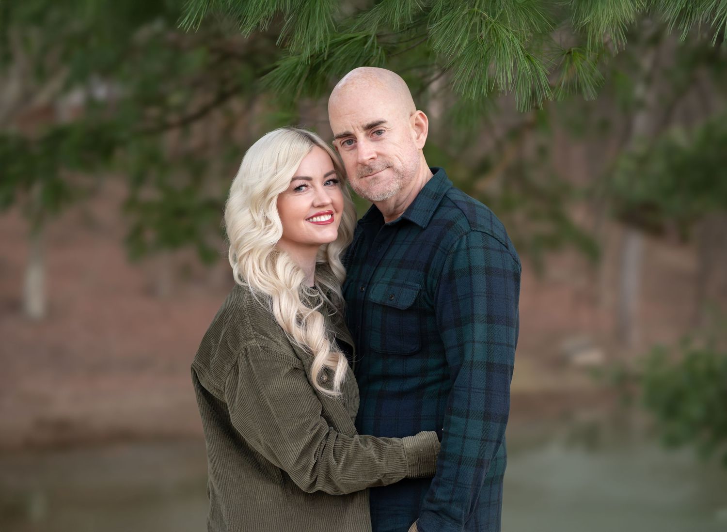a husband and wife embrace and smile for a Christmas card picture with evergreen trees and a lake in the background