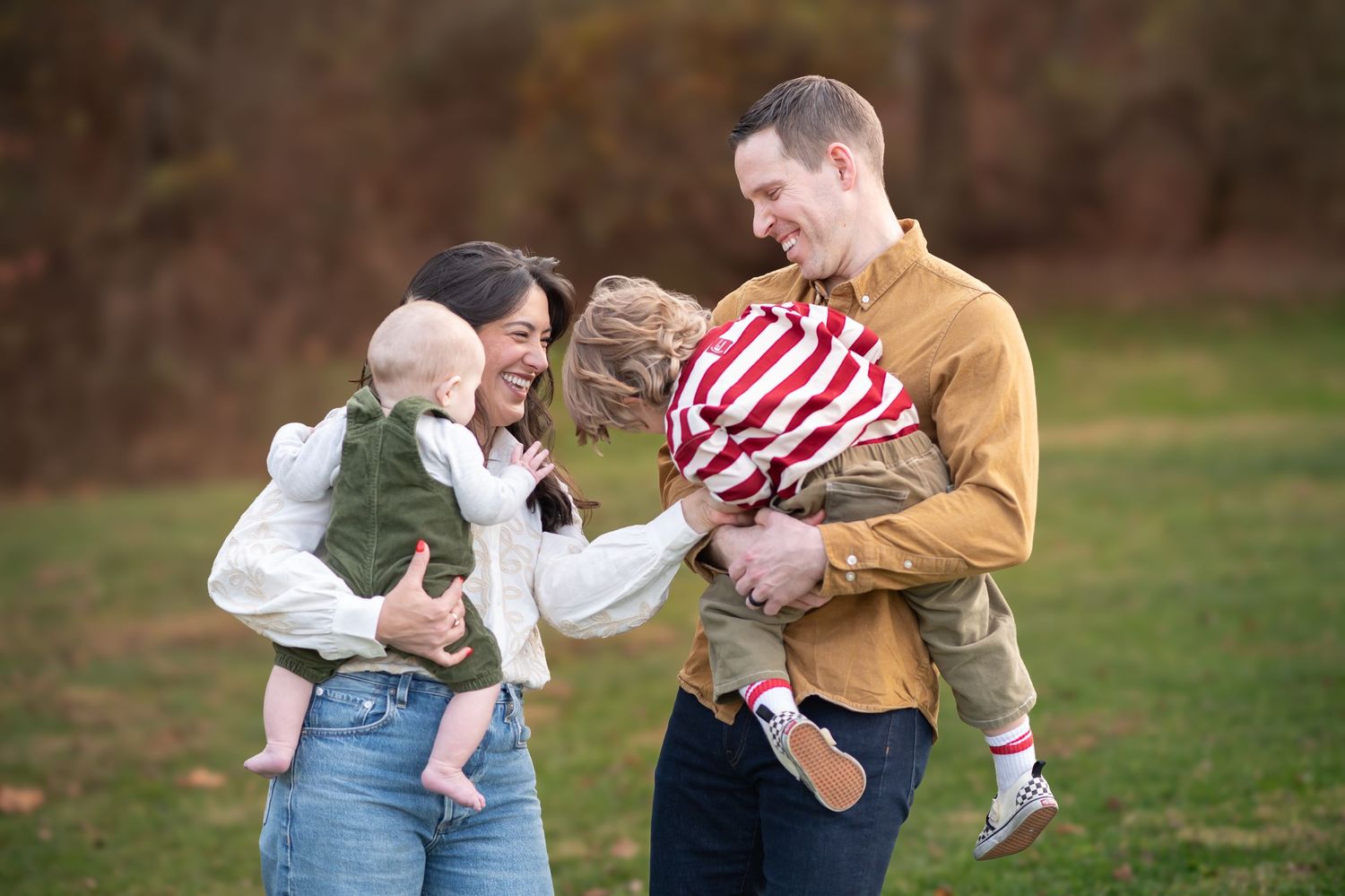 a young couple holding their two small boys tickles them and laughs together, with beautiful fall scenery in the background