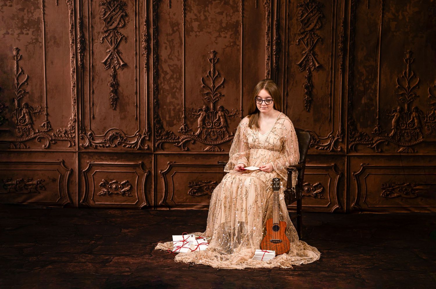 a lovely young woman with long brown hair in a shimmering gold floor length gown sits in a leather chair with letters scattered around her