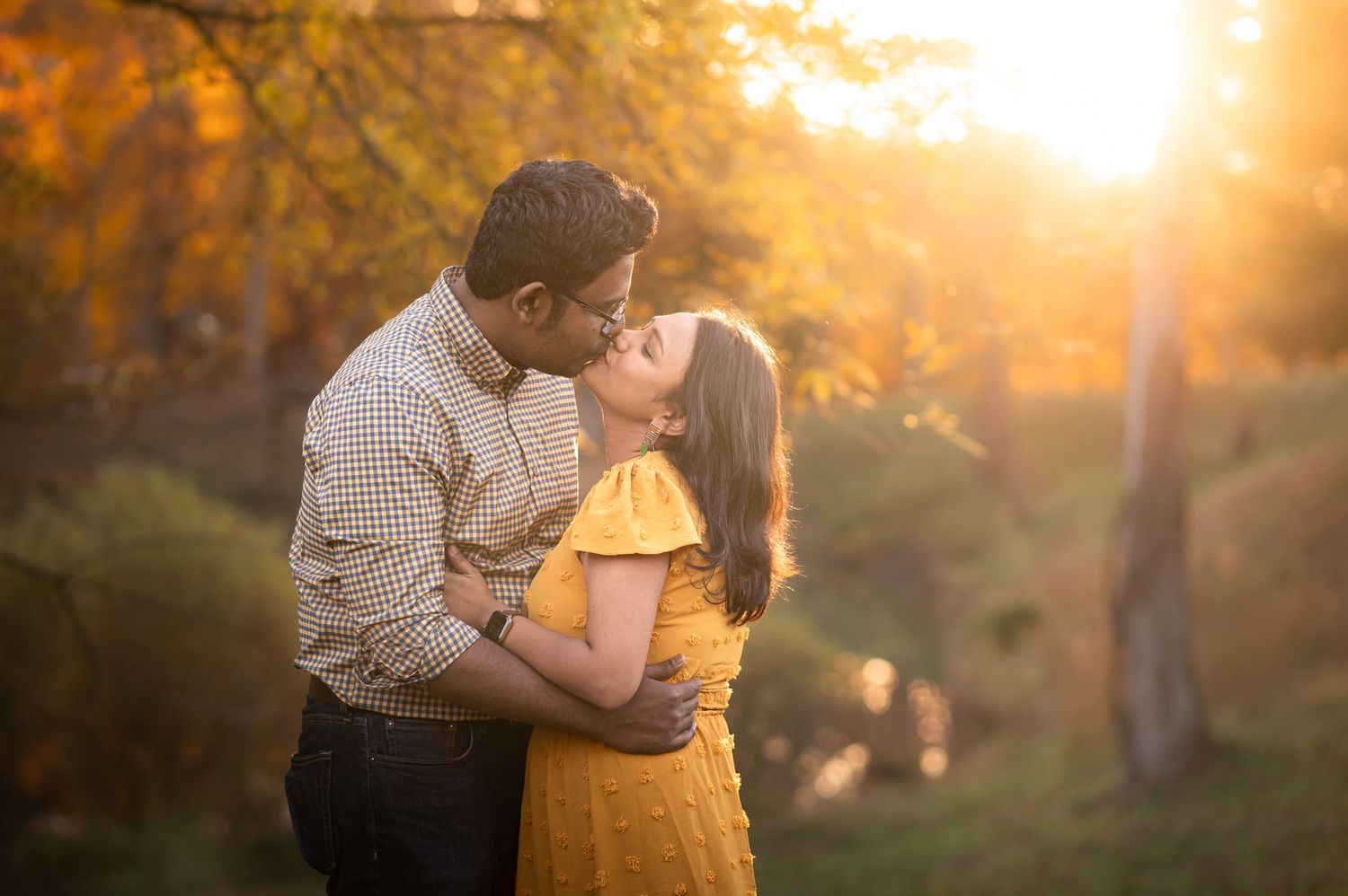 a husband and wife kiss for a portrait in front of a beautiful glowing golden backdrop of autumn foliage lit by the setting sun