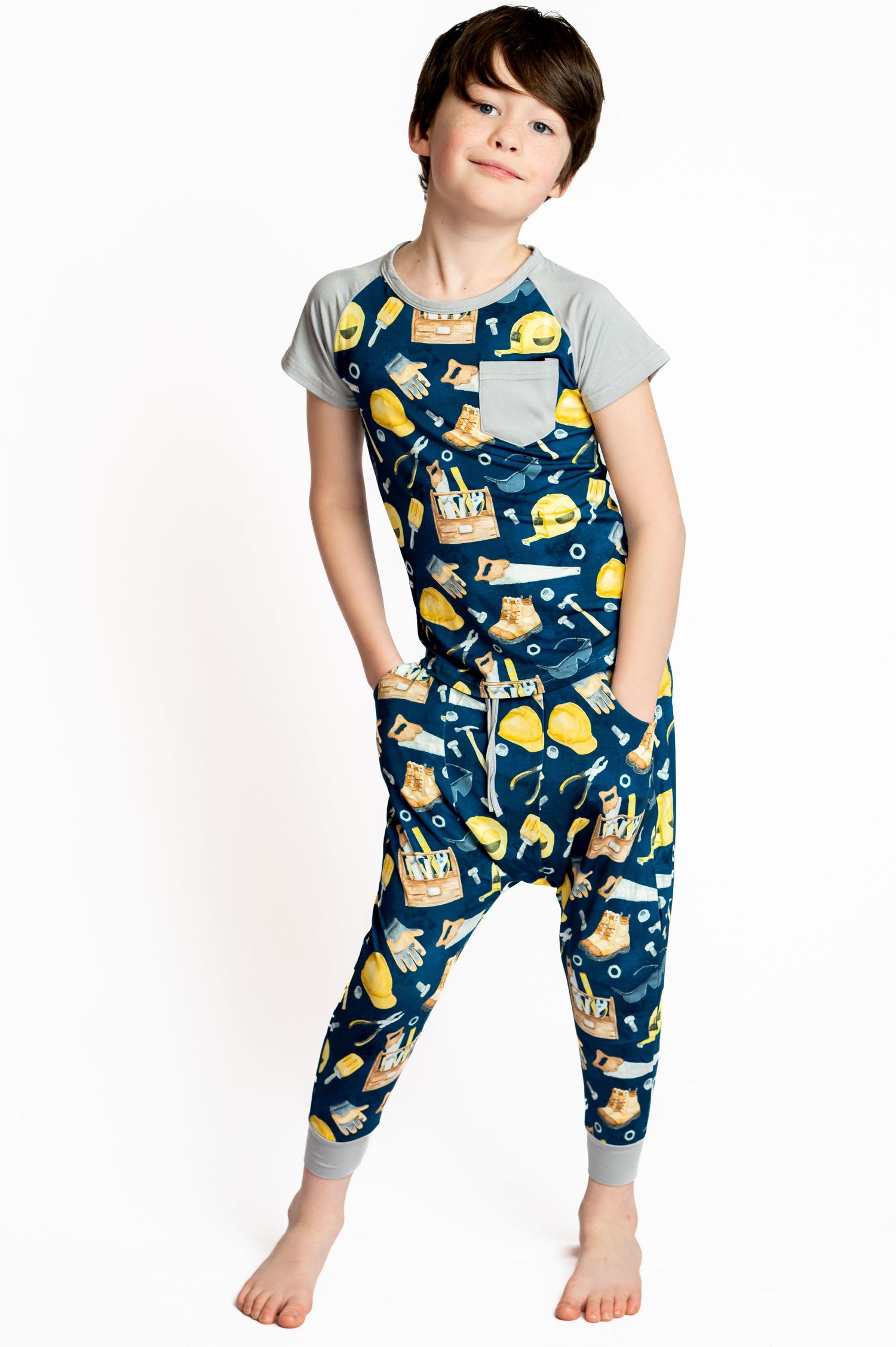 A boy models navy blue tool-print jumpsuit with gray raglan sleeves and cuffed ankles, posing barefoot on a white studio backdrop.