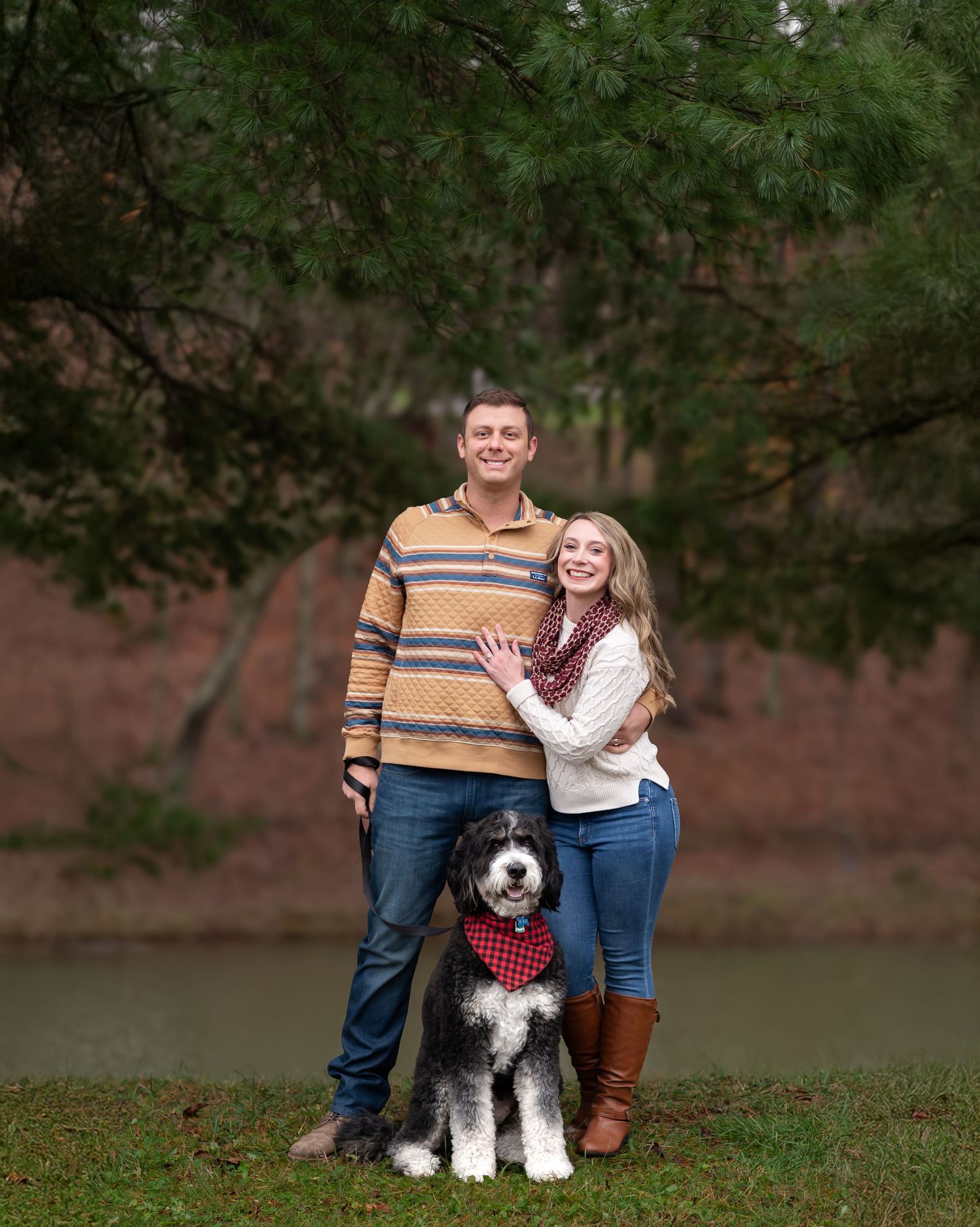 Couple smiling outdoors with their black and white dog wearing a red bandana, standing near a pond under pine trees.