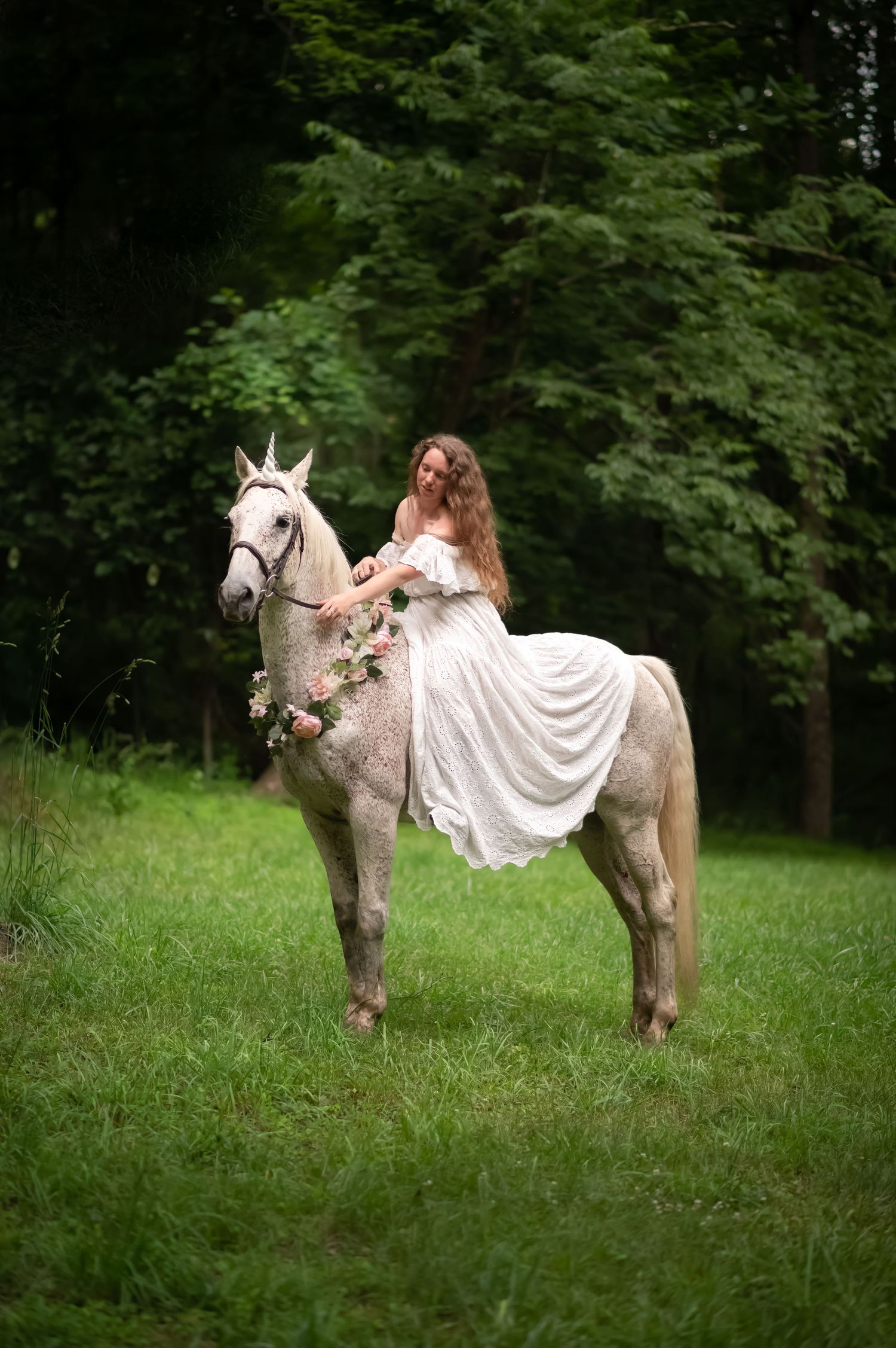 a young woman in a long white gown sits on the back of a white unicorn in a deep green grassy clearing with woods all around