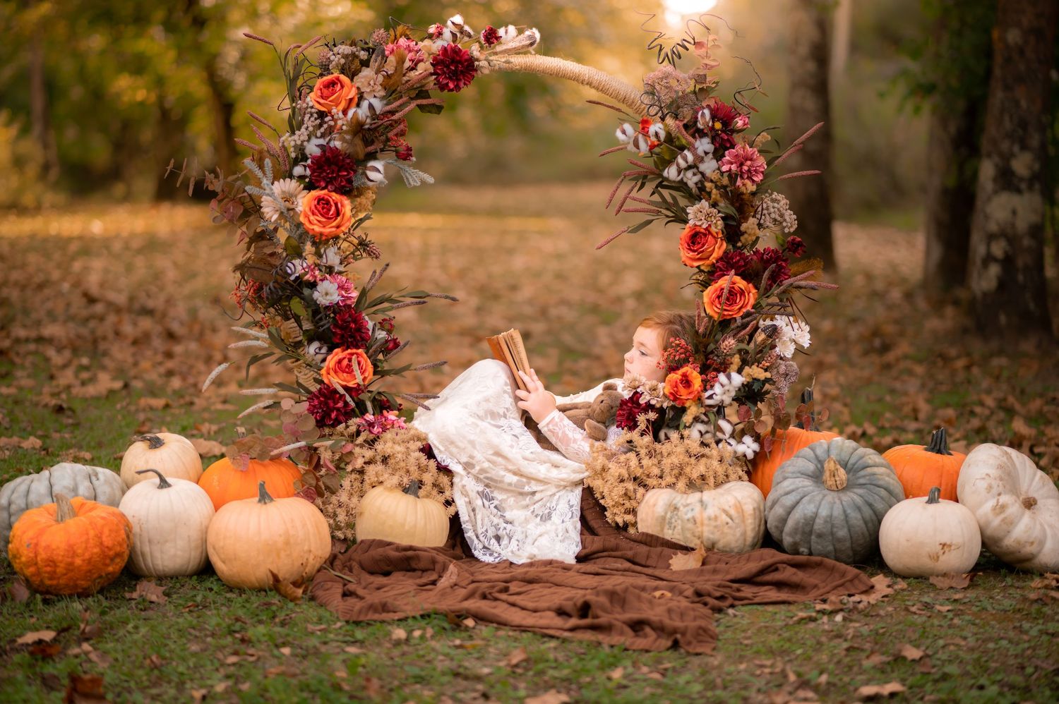 a little red headed girl in a white lace dress lounges in an arch of autumn flowers surrounded by pumpkins and reads a book