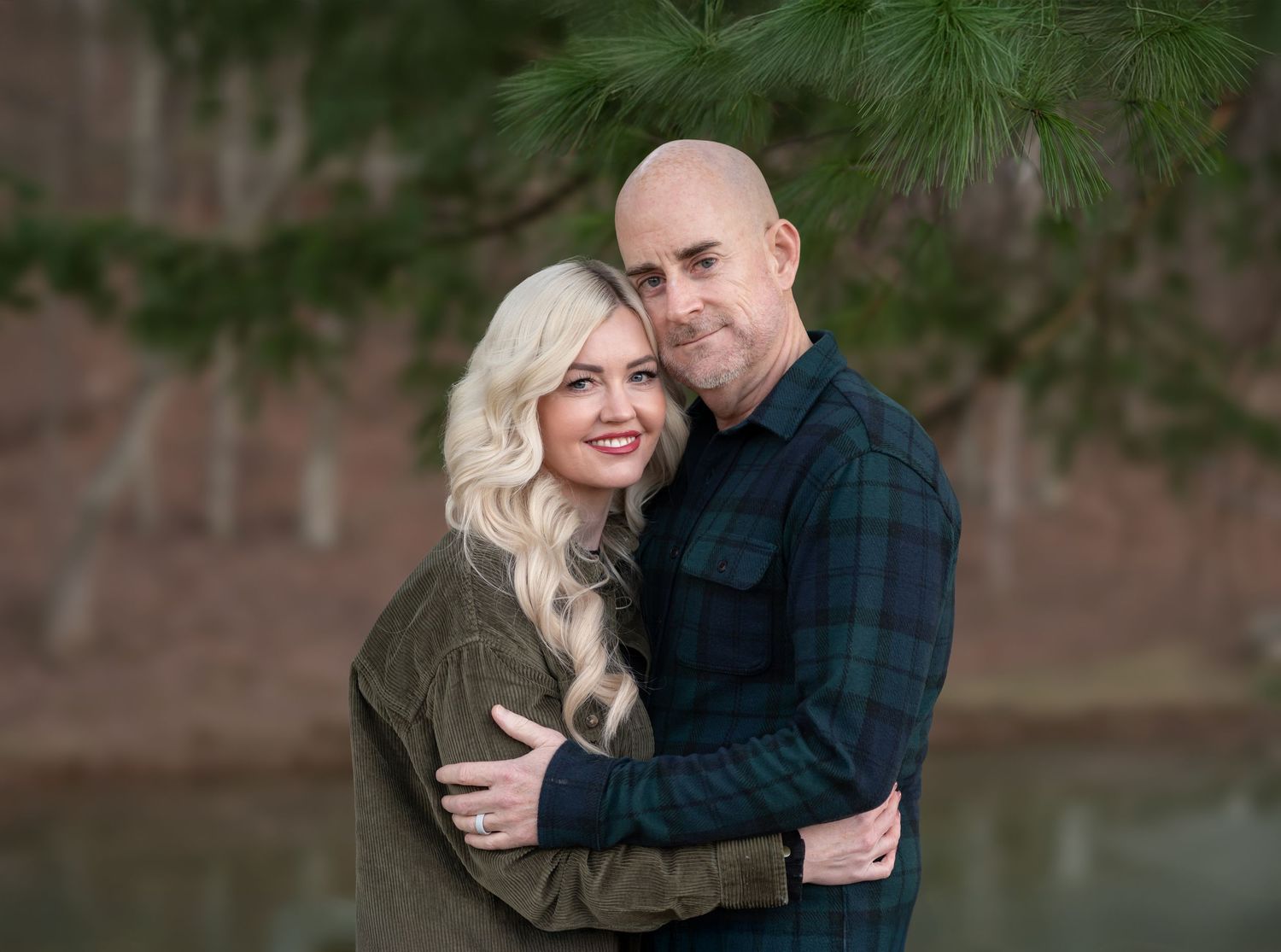a blonde woman and her husband pose for a Christmas portrait outside, surrounded by evergreen trees