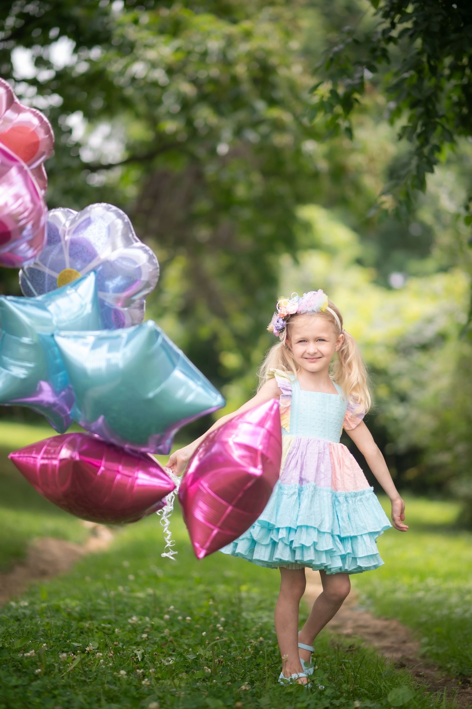 a little girl in a pastel rainbow dress holds a large bunch of pink and blue balloons and walks through the grass with trees behind