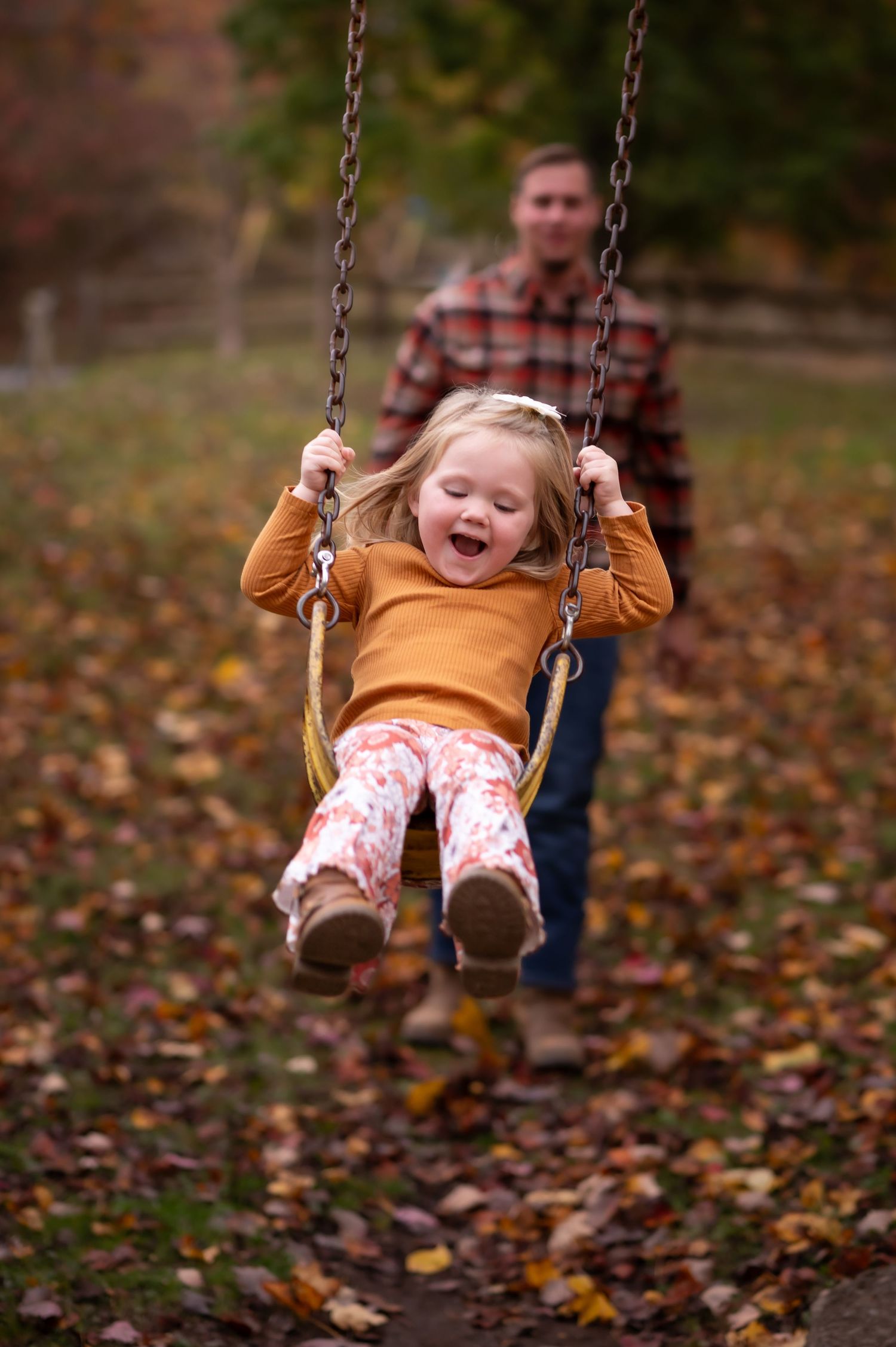 a little girl swings high on a park swing and laughs, while her father stands blurred in the background, pushing her swing