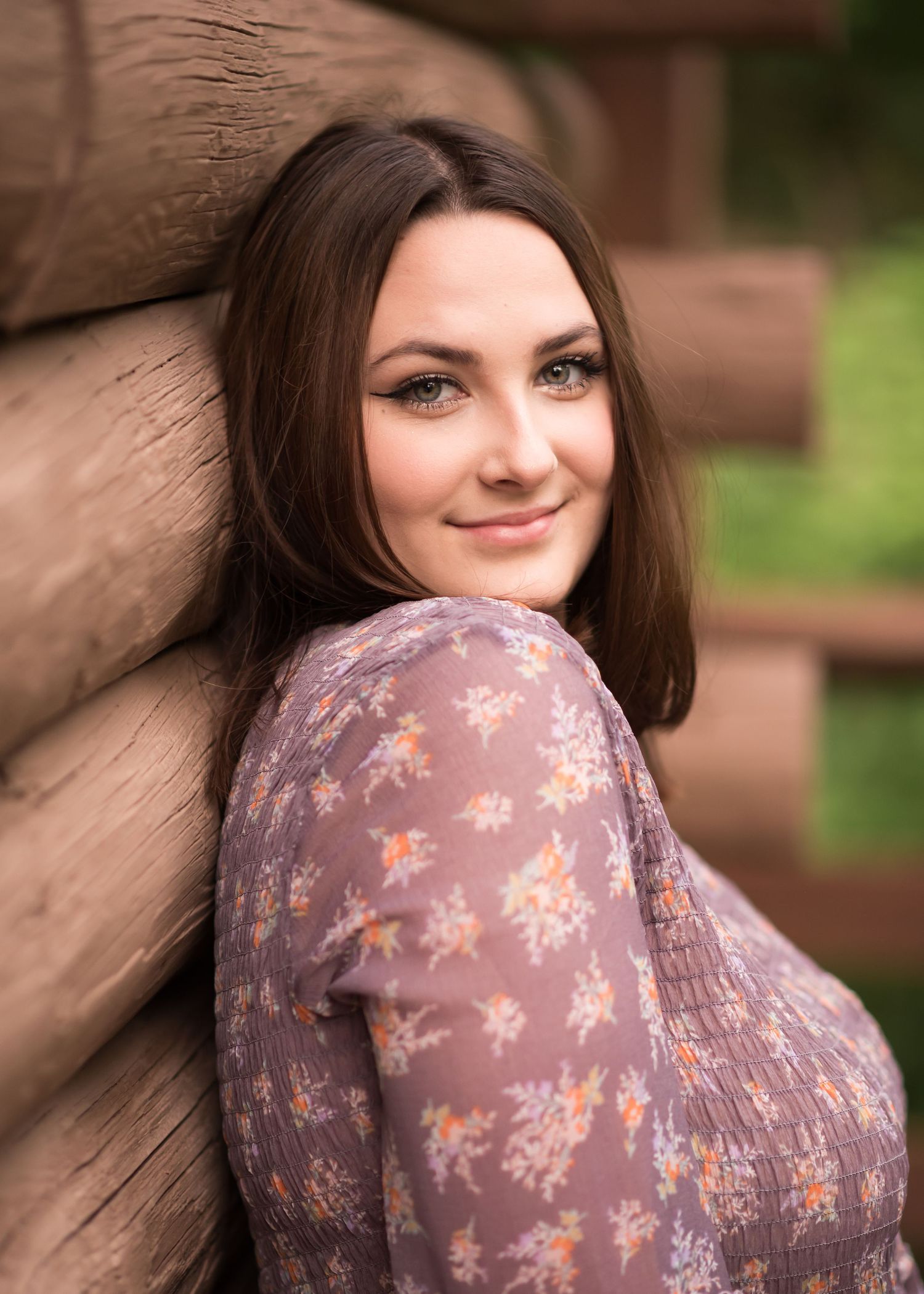 a dark haired senior student in a purple dress leans against the wall of a log cabin and smiles at the camera for a closeup portrait