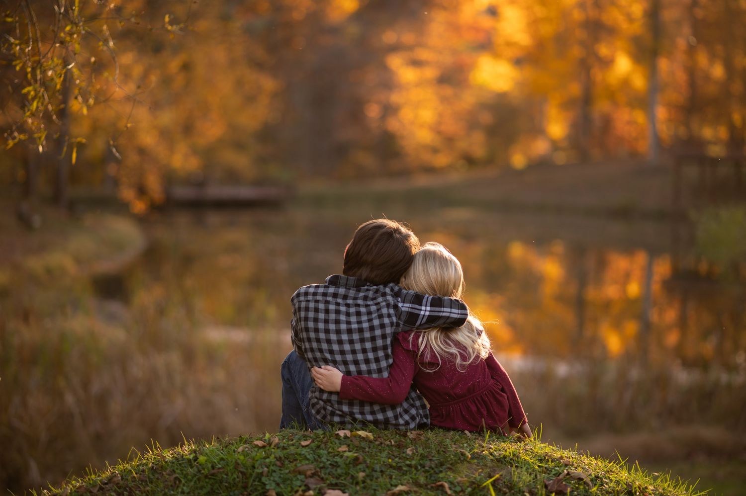 two siblings sit together looking over a pond, with brilliant orange and gold autumn foliage in the background and reflected in the pond