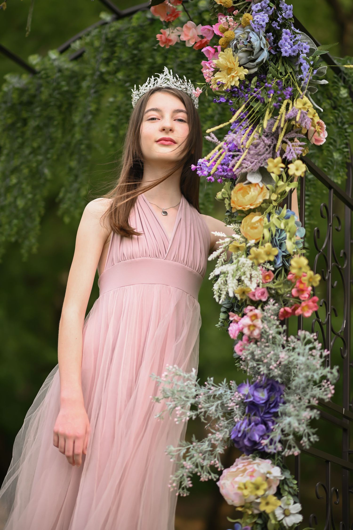 princess portrait of a young woman in a long pink gown with a tiara, standing by a tall floral arch