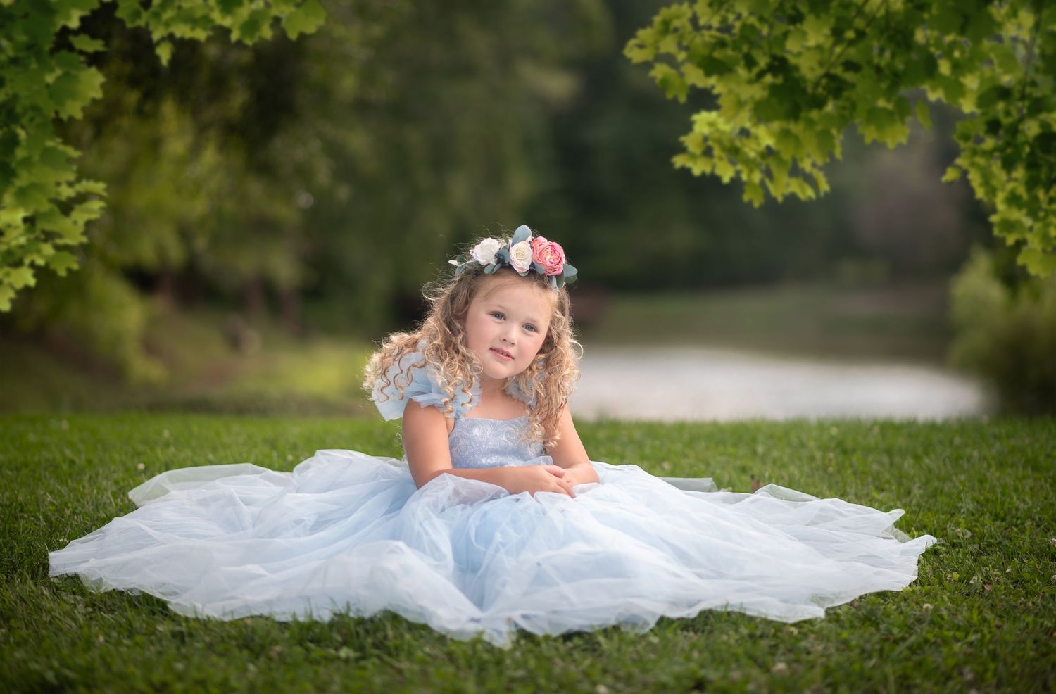 a lovely little girl in a fluffy blue dress and a floral crown sits in the green grass with a pond and trees behind her.