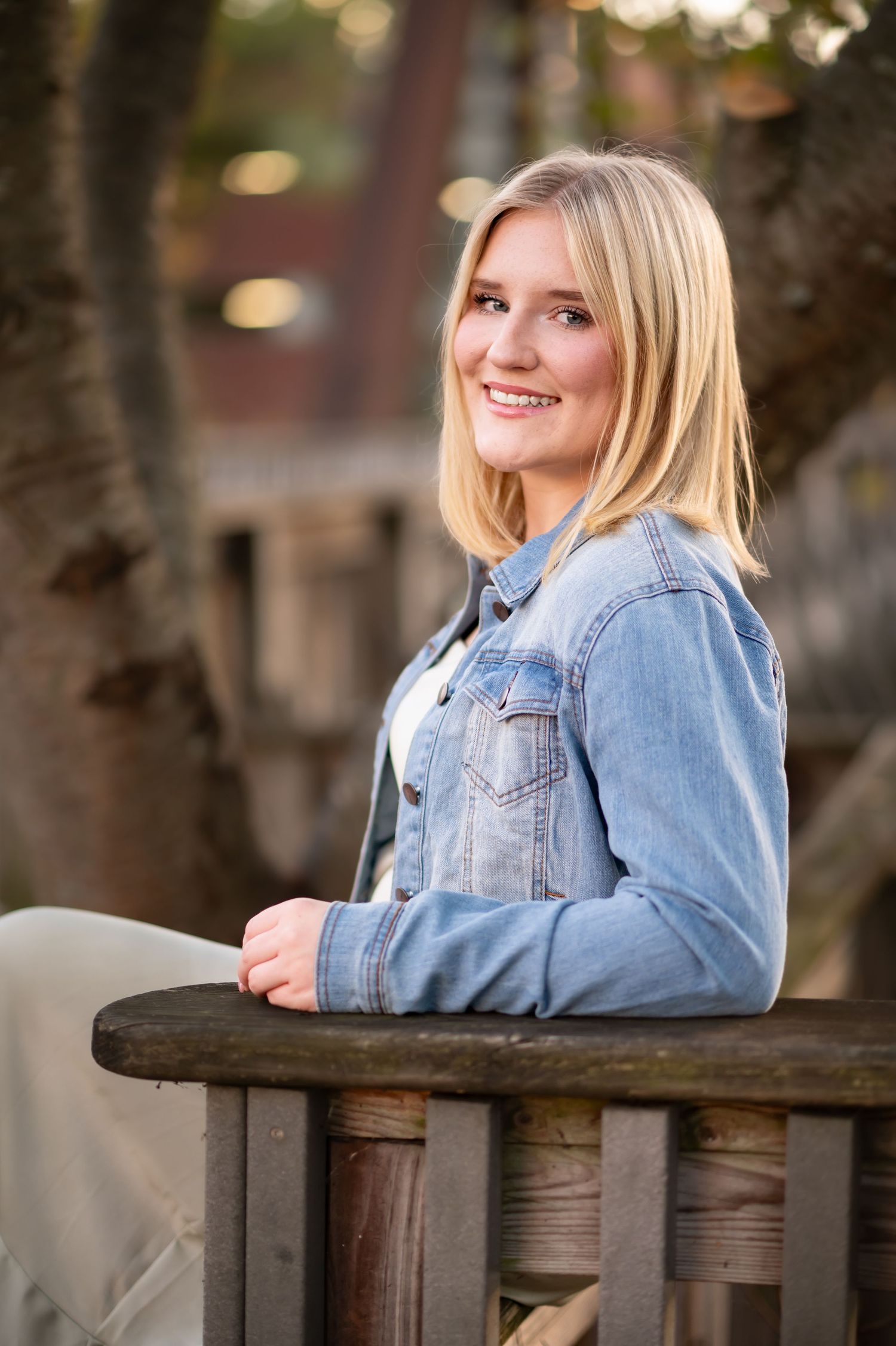 senior portrait of a young blonde woman in a jean jacket, posing with a smile beside a wooden railing