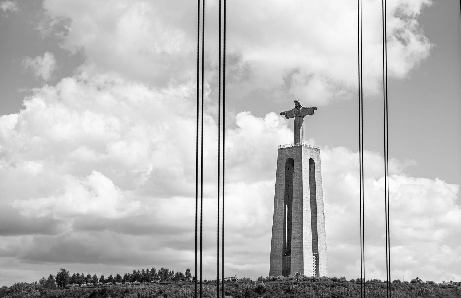 Black and white photo of Cristo Rei statue in Lisbon, Portugal, standing tall against a cloudy sky.