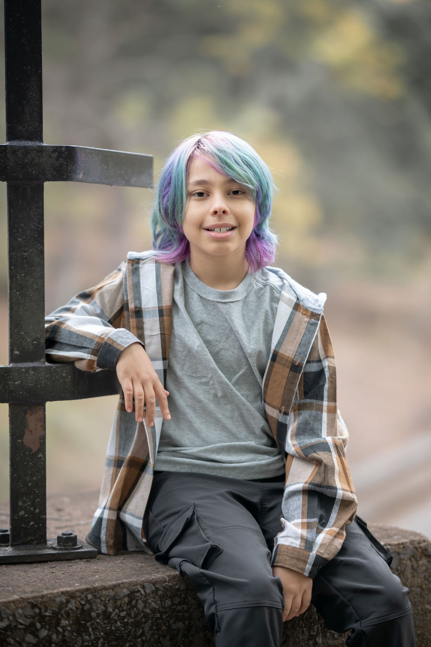 A young boy with colorful purple and blue hair sits smiling outdoors, wearing a plaid shirt and gray top.