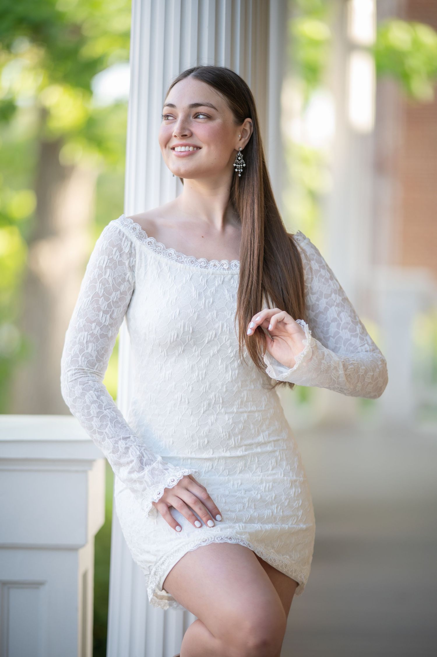 a graduate student in a white dress leans against a white pillar and smiles