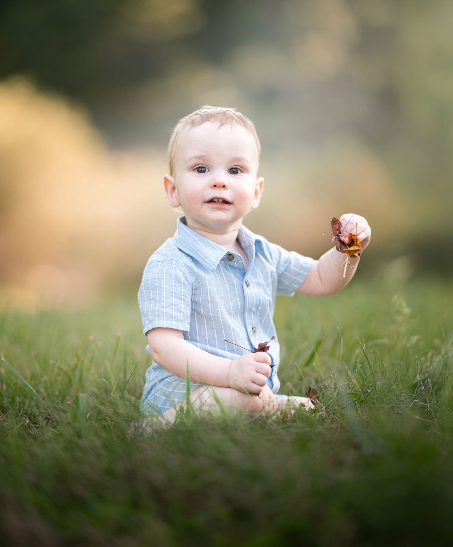 a little blonde boy in a blue romper sits in the grass holding a leaf, and smiles at the camera during an outdoor first birthday session