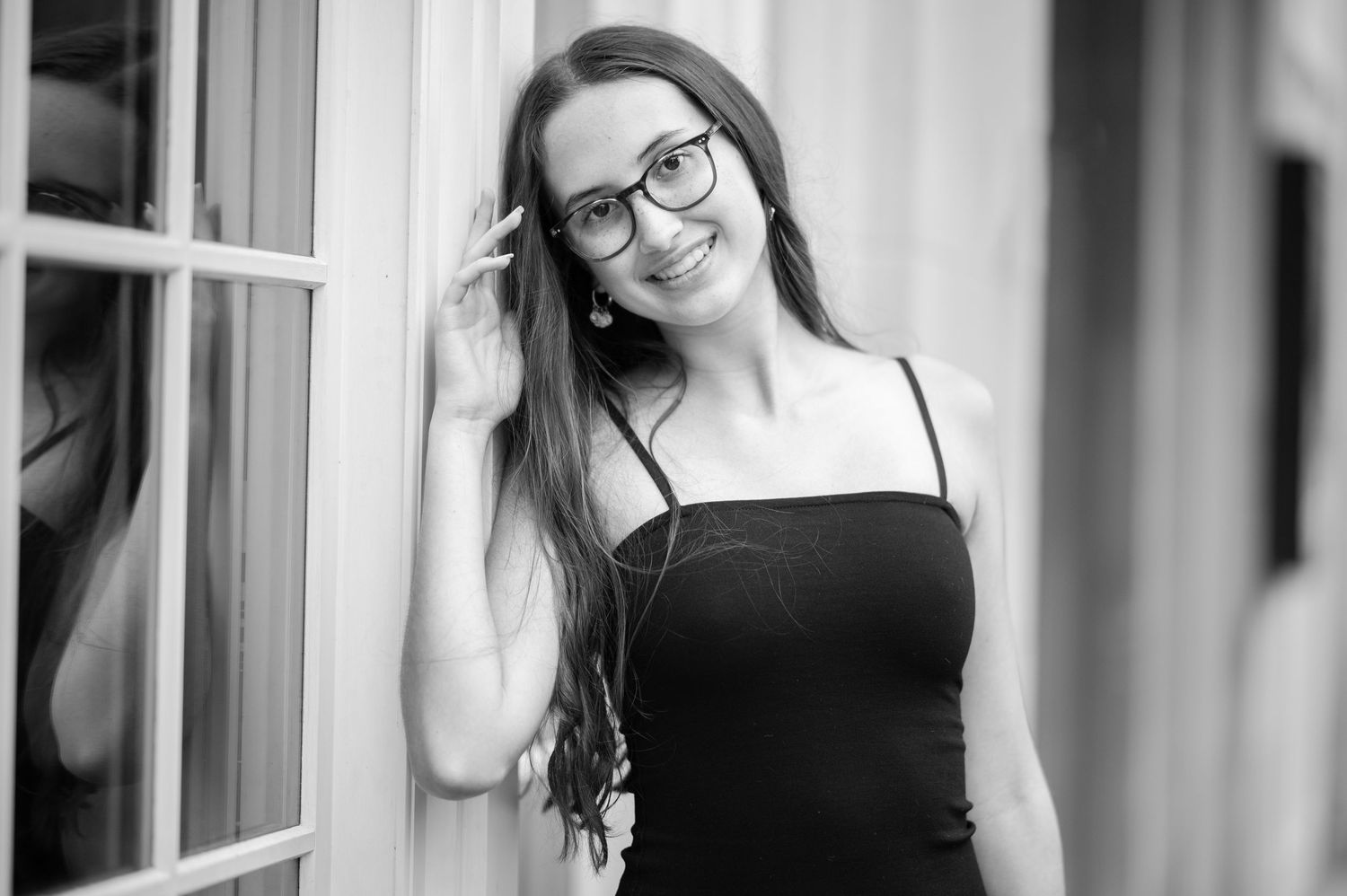 black and white portrait of a lovely senior student with long dark hair and glasses leaning against a wall of windows in downtown Morgantown