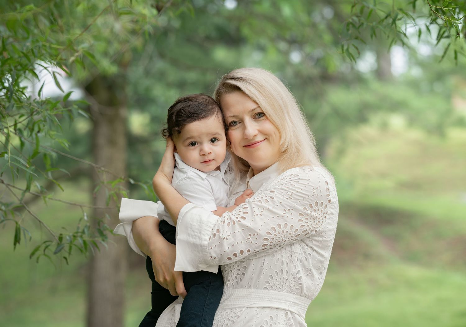 a blonde mother dressed in white holds her little son with their faces close together, and sunlit green trees in the background