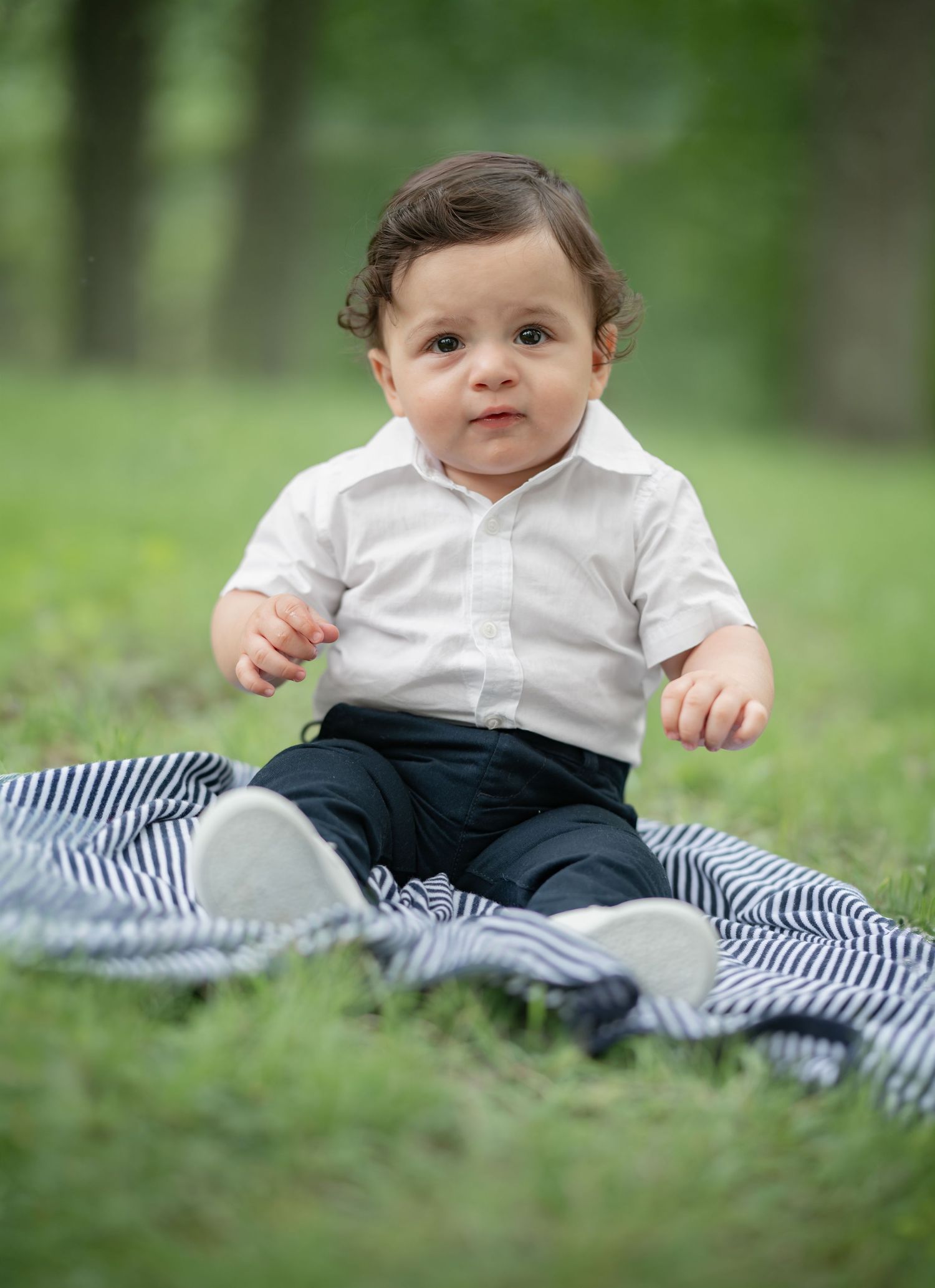 a little boy with dark hair and dark eyes in a white shirt sits on a blanket in the grass for a portrait in Morgantown, WV
