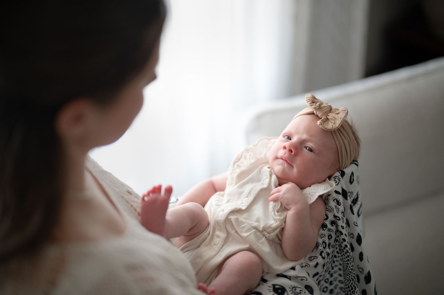 A mother tenderly holds her newborn baby dressed in a white lace outfit with a bow headband near a bright window.