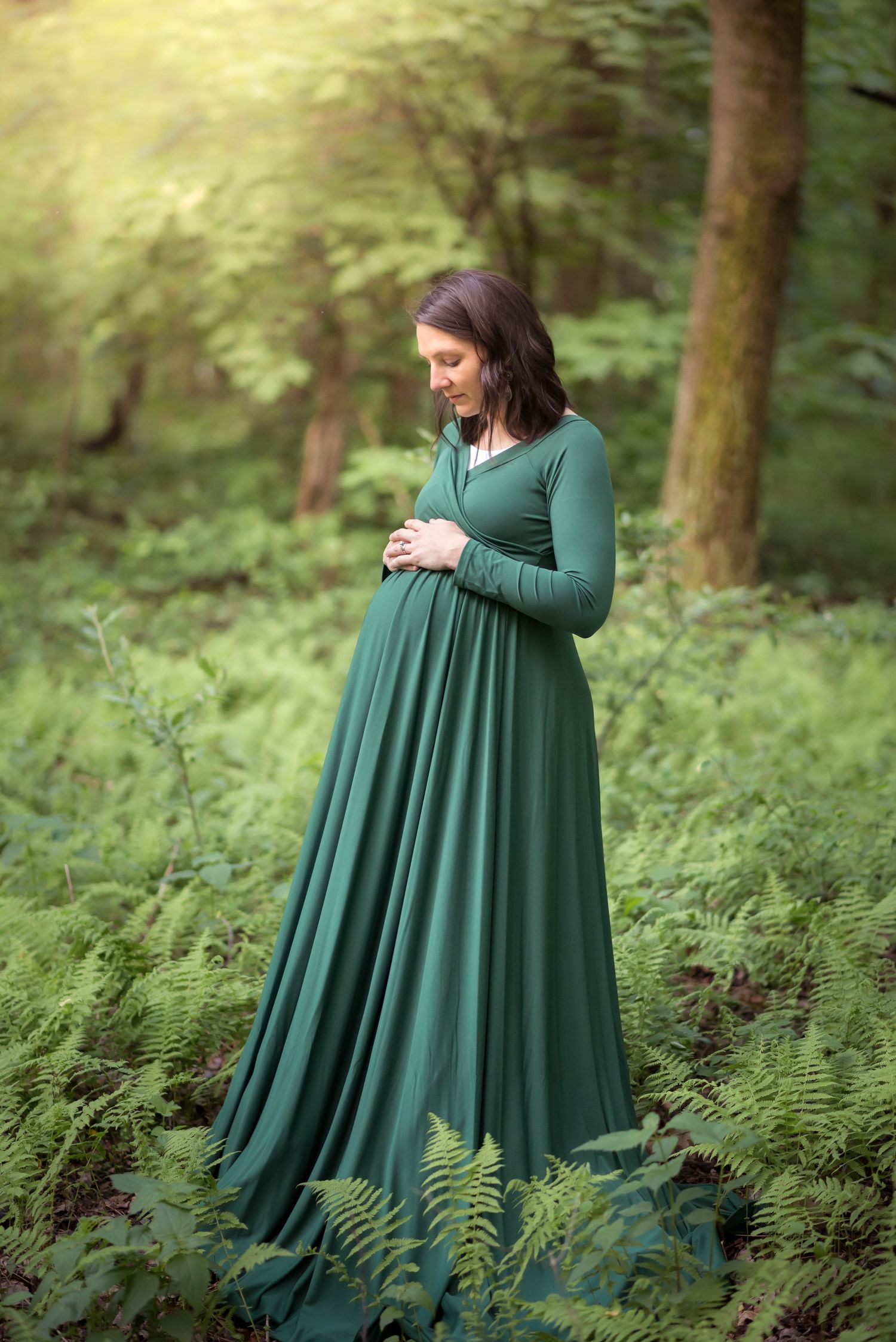 a dark haired pregnant woman in a long green dress stands in the forest surrounded by tall ferns