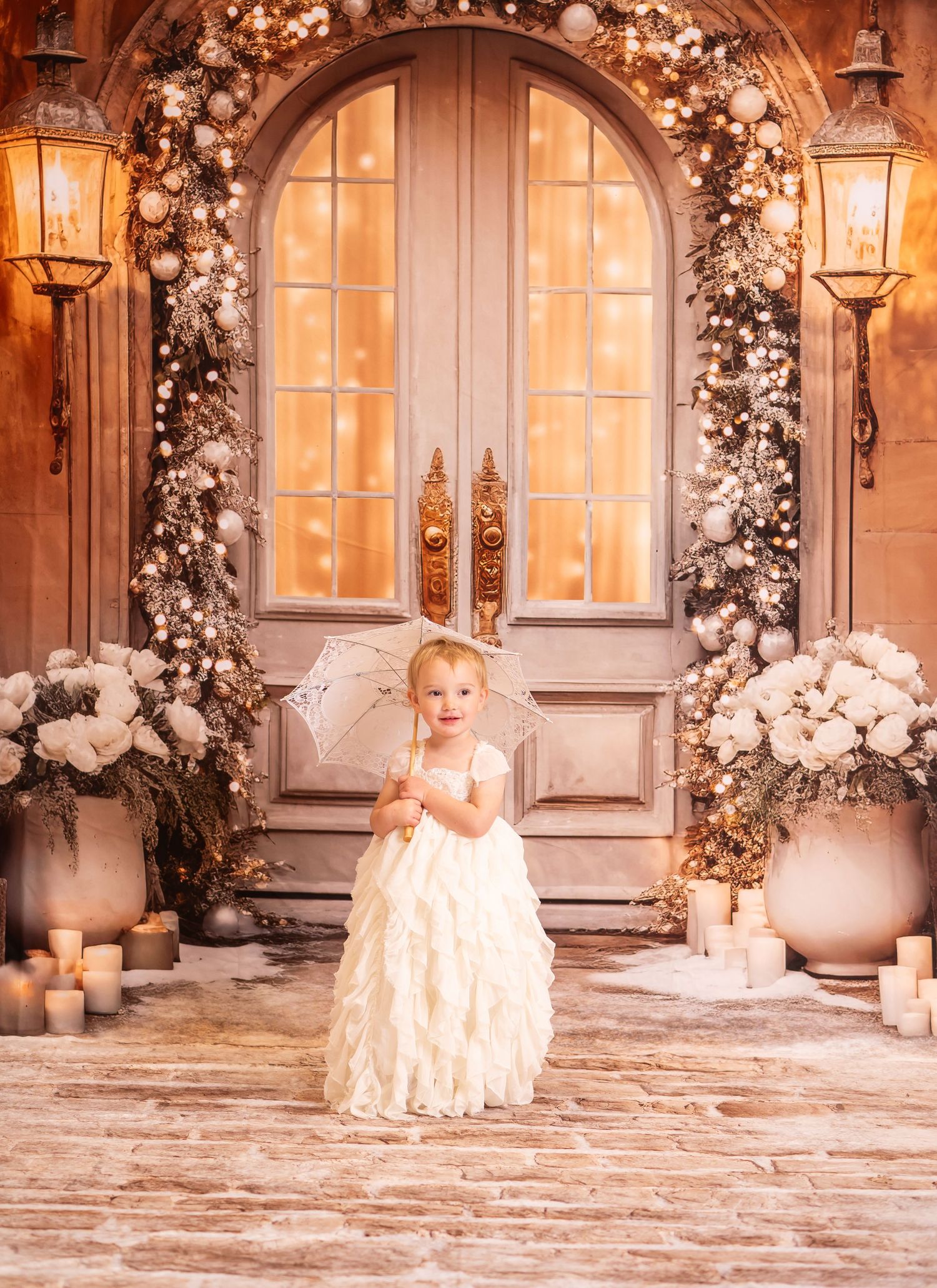 a toddler girl in a frilly white gown holds a parasol and poses on a brick street in front of a doorway decorated with white and gold lights