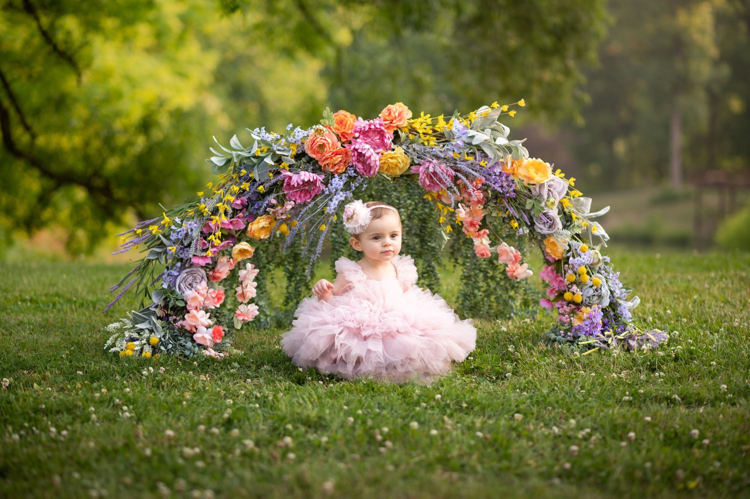 a sweet baby girl in a pink tutu dress sits under a floral arch in the green grass with green trees in the background