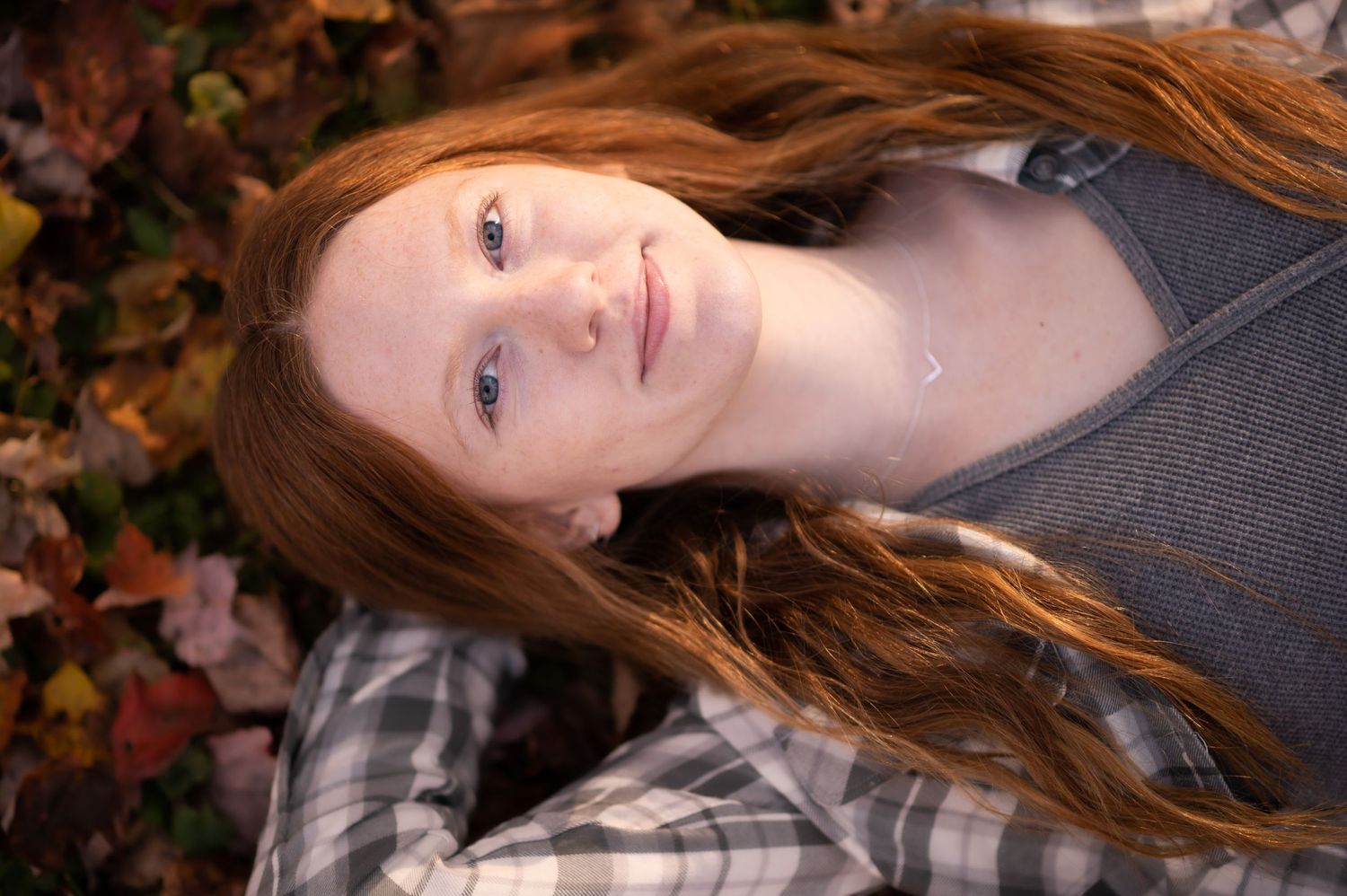 portrait of a young red-haired woman in plaid lying on her back on the ground among fall leaves, looking up into the camera with a smile