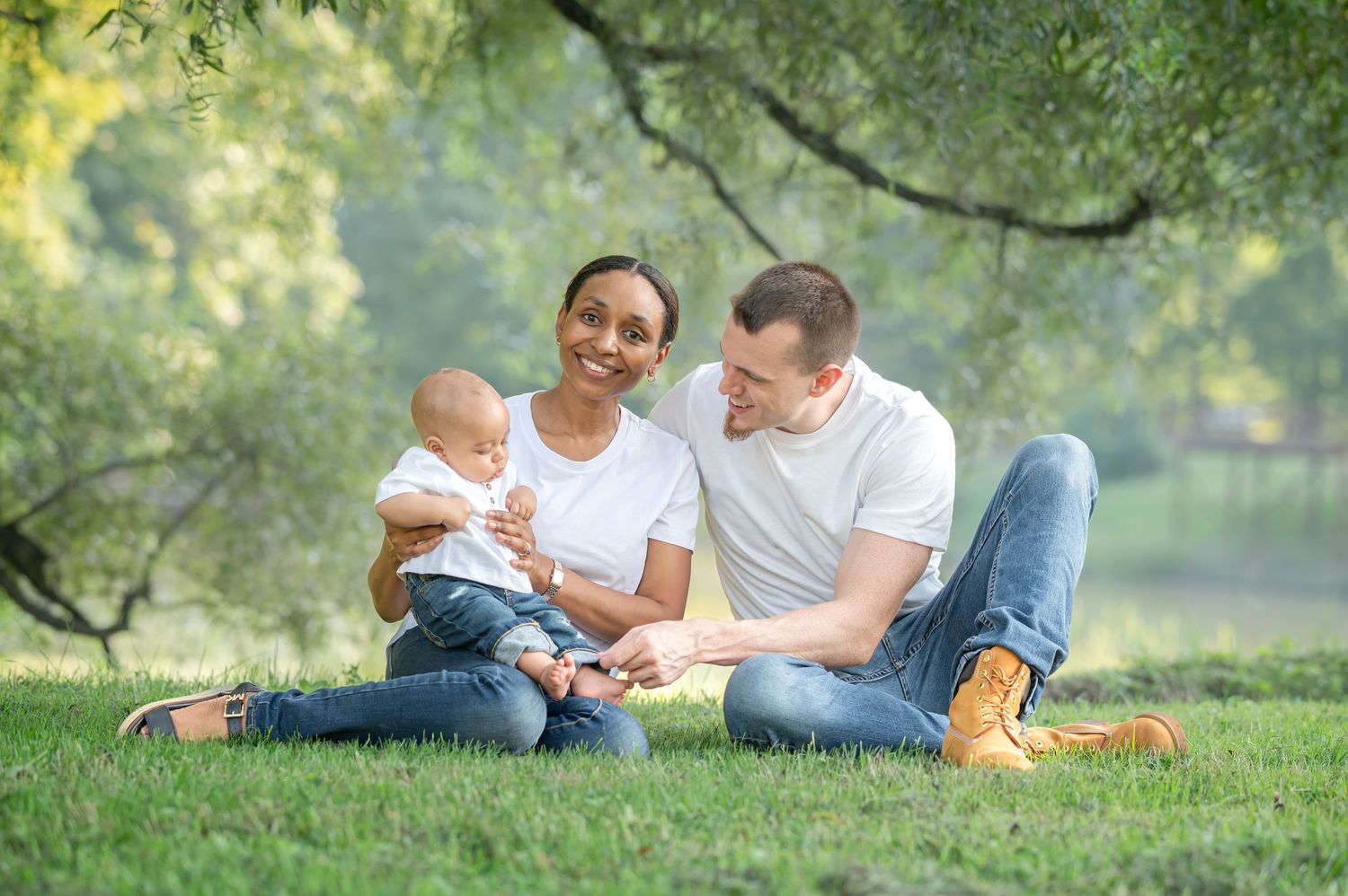 a young couple in blue jeans and white tshirts sit in their grass with their baby boy, with soft green trees in the background