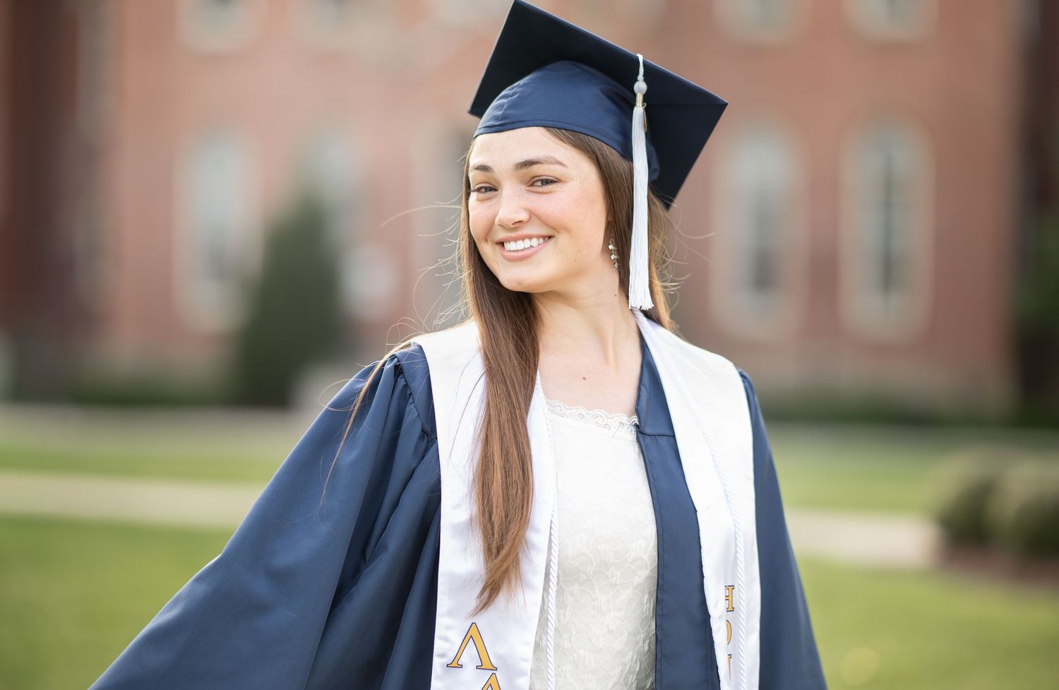 closeup of a WVU graduate student in her blue cap and gown, smiling in front of Woodburn Hall on WVU's campus