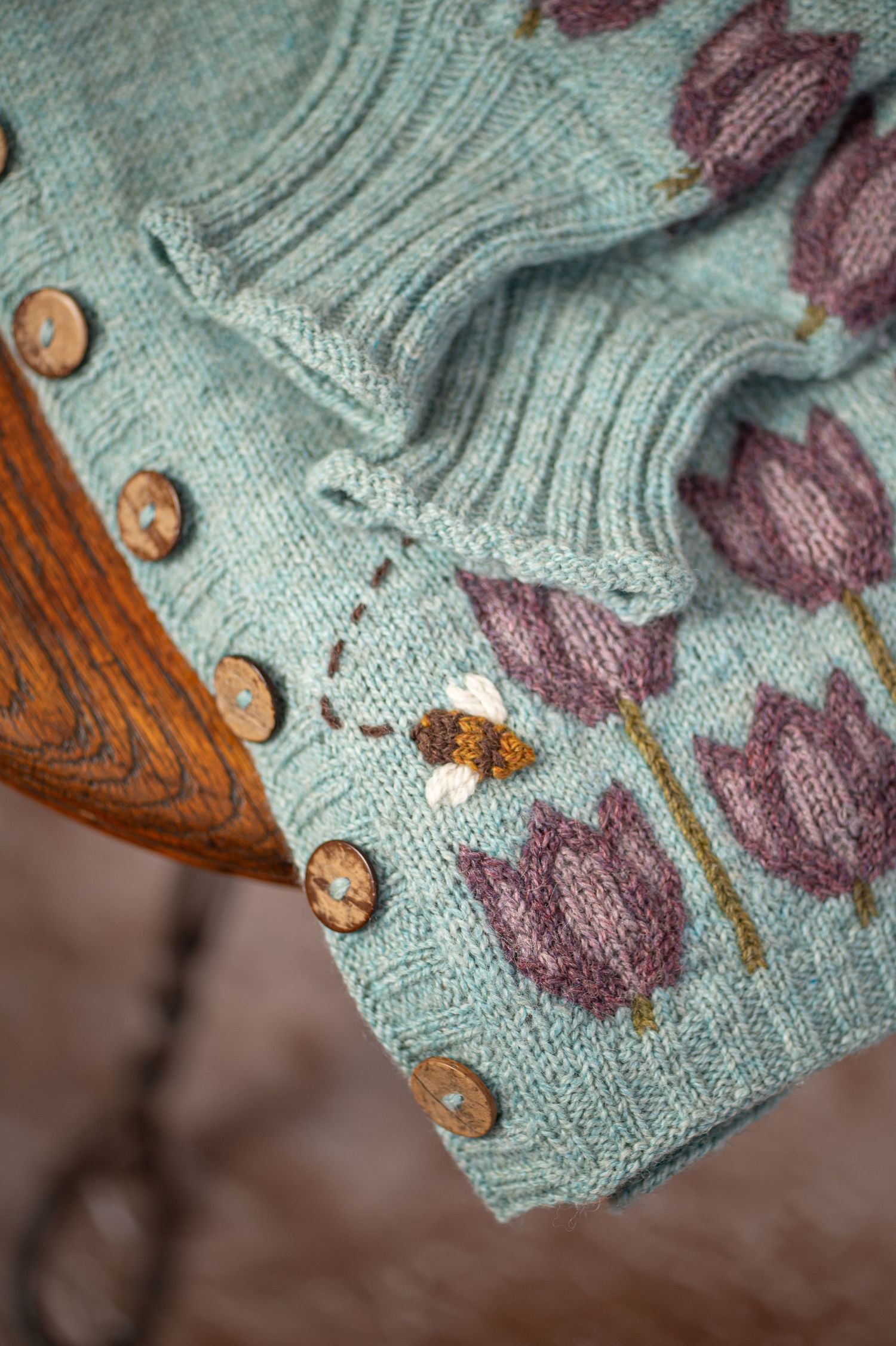 a detail shot of an intricate hand knitted sweater by Rountree Stitchery, featuring a design of purple tulips worked into soft teal yarn