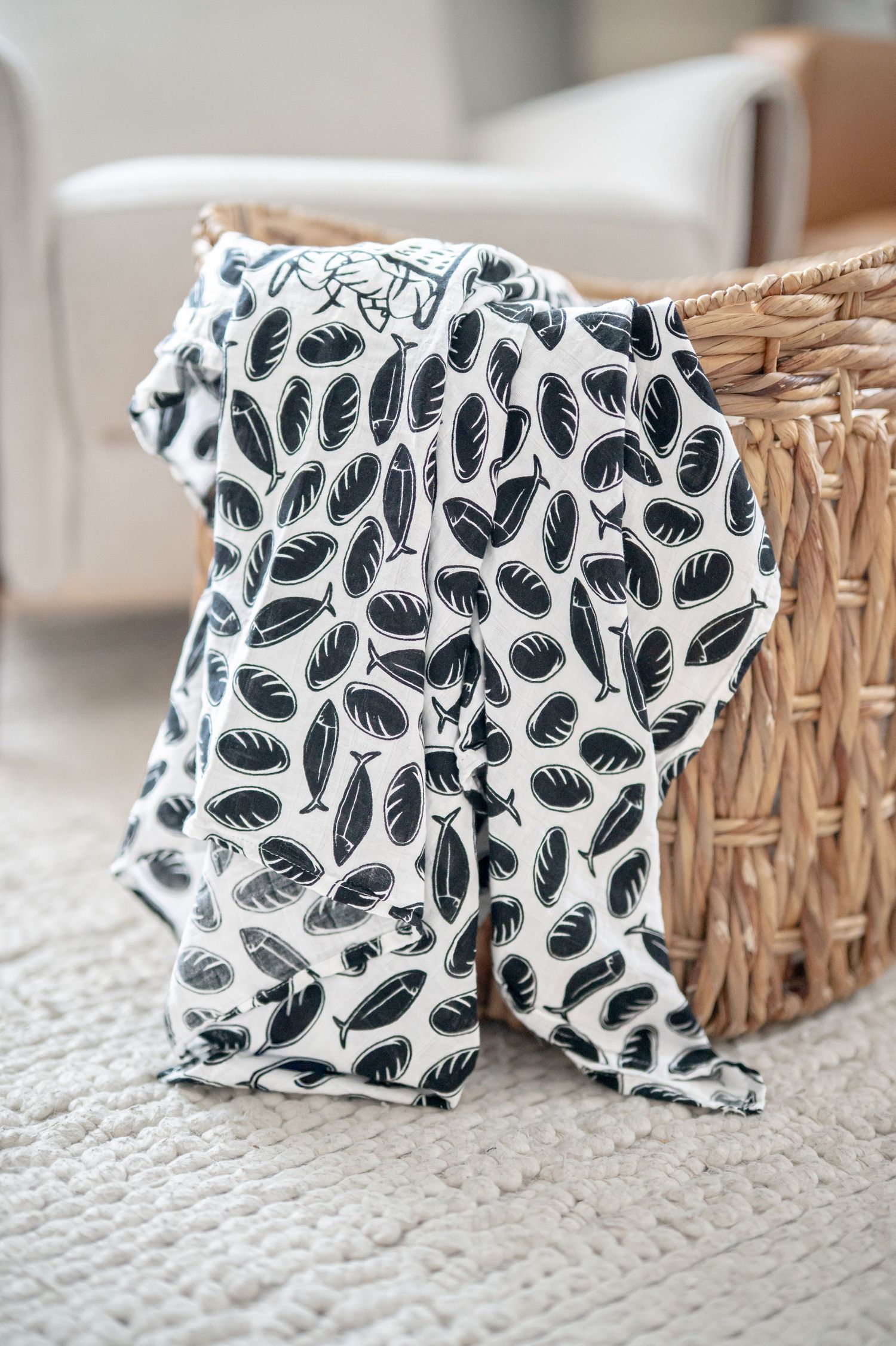 Commercial product image of a white muslin swaddle blanket with black loaves and fishes pattern draped over a wicker basket on a white rug.