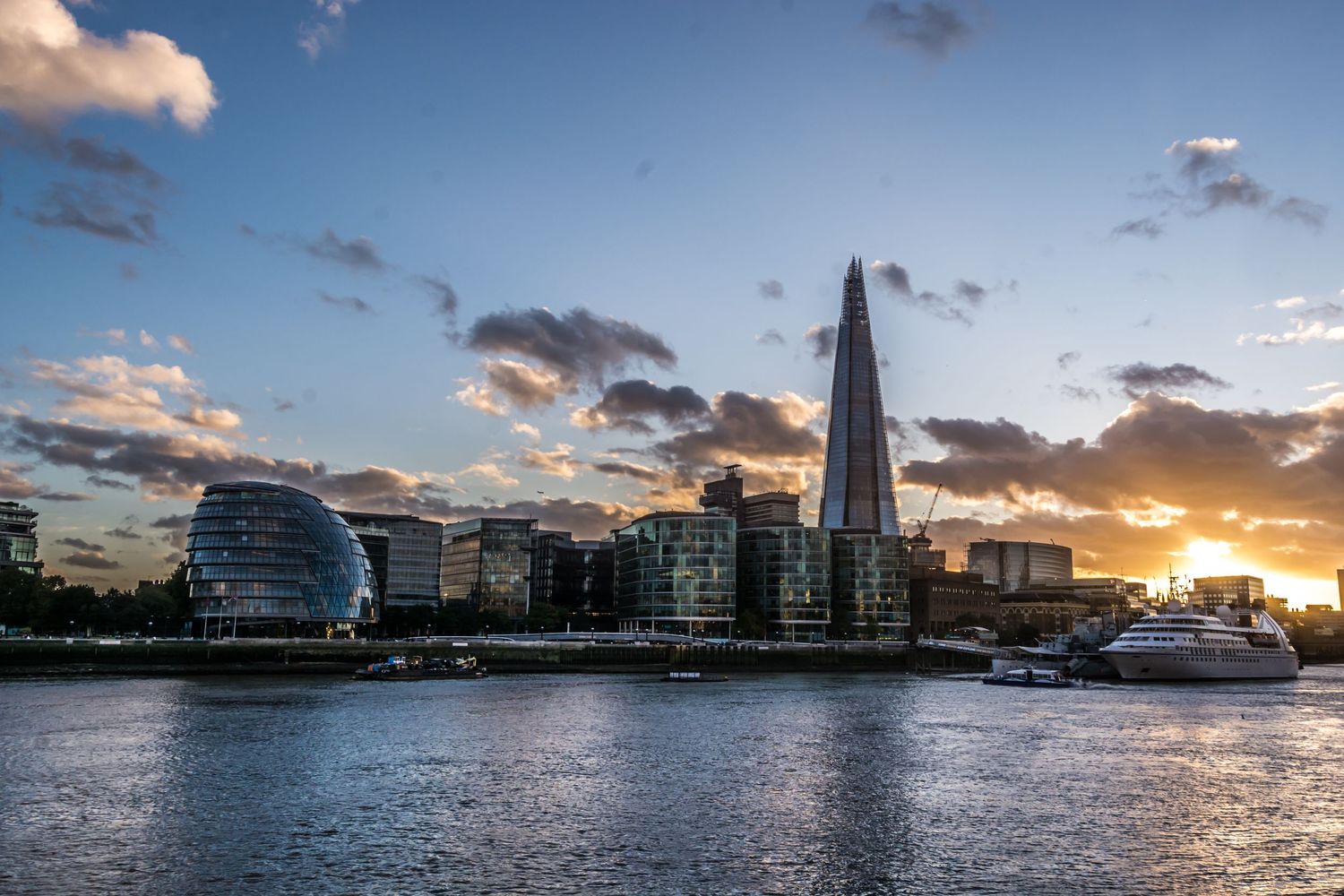 London skyline at sunset featuring The Shard skyscraper and City Hall reflecting on the Thames River.