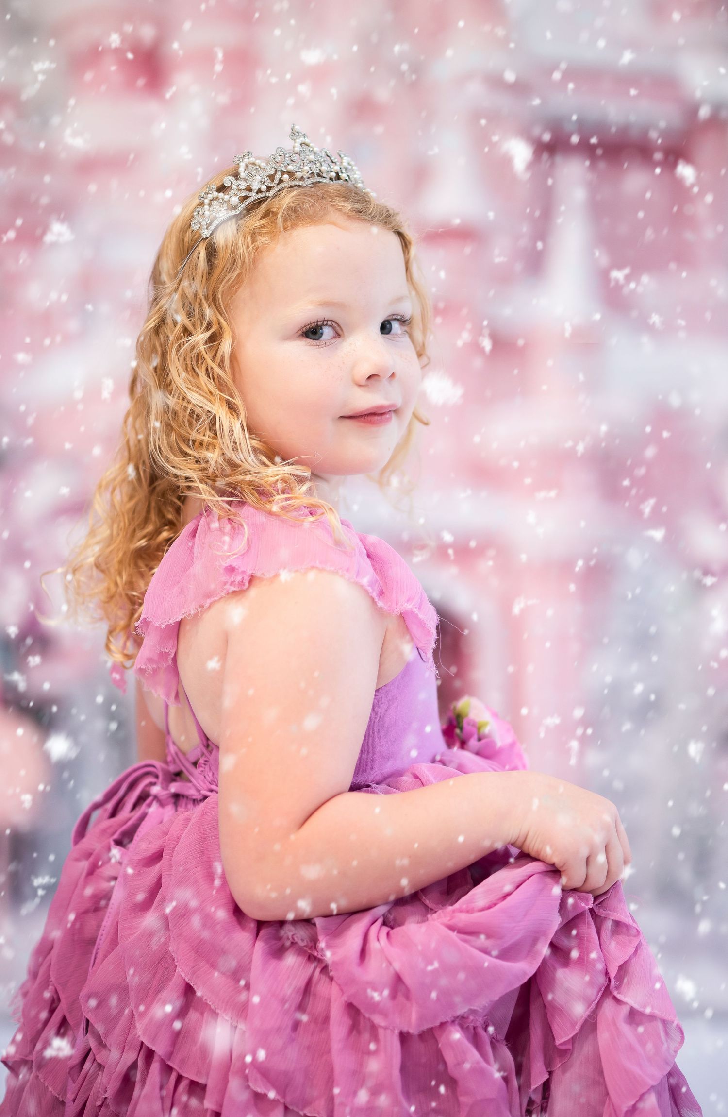 a little girl in a frilly pink gown stands in the snow with a pink candy Christmas castle behind her.