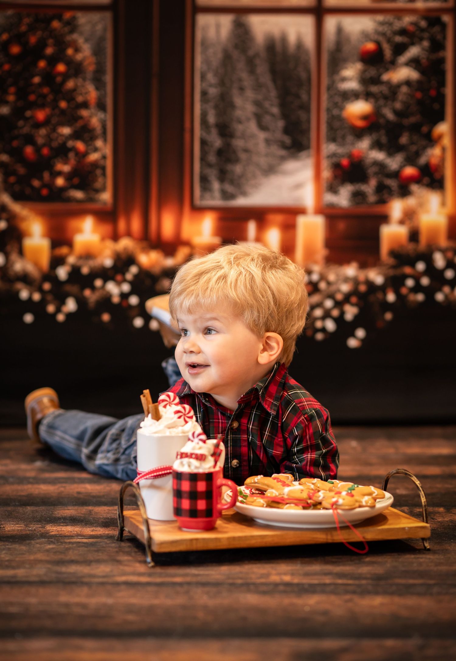 a little blonde boy lies on a wood floor on his belly with a tray of hot cocoa and cookies in front of him, and Christmas trees and candles