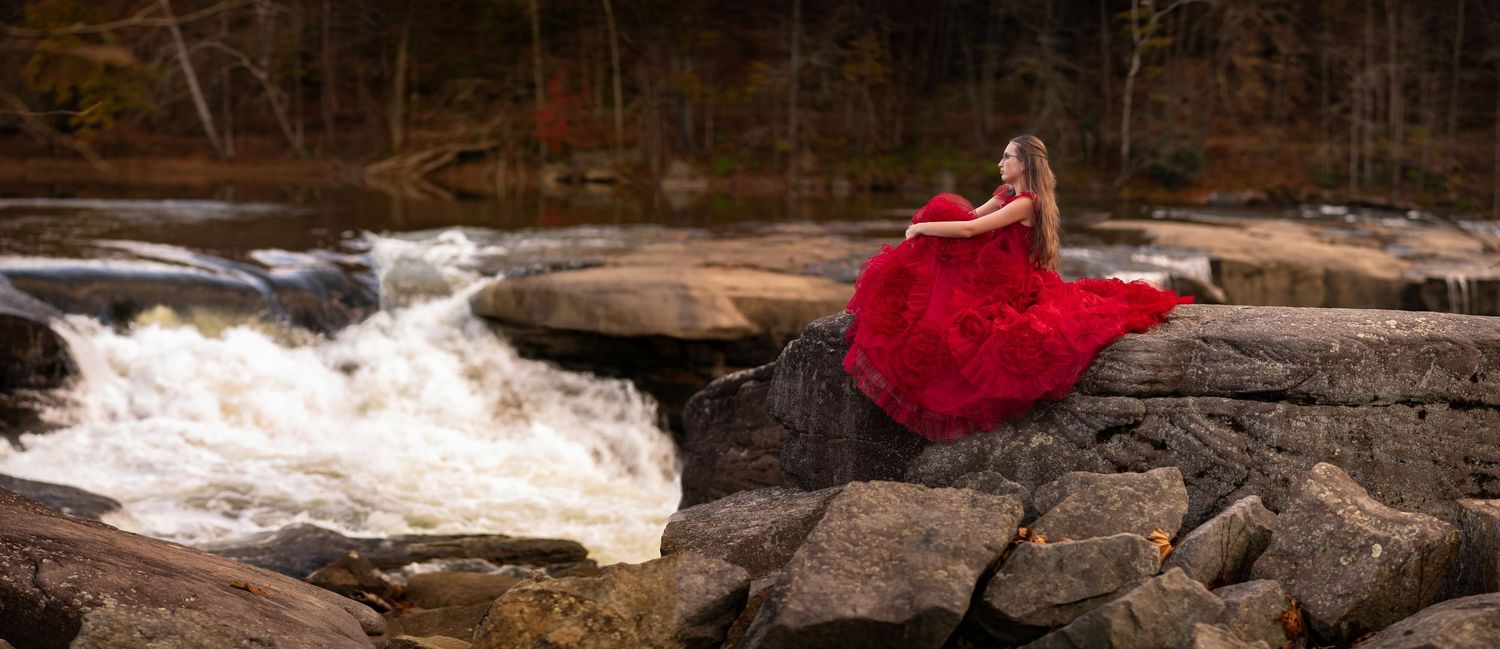 a young woman with long brown hair in a dramatic red dress sits on a large rock overlooking a rushing waterfall, in Valley Falls State Park