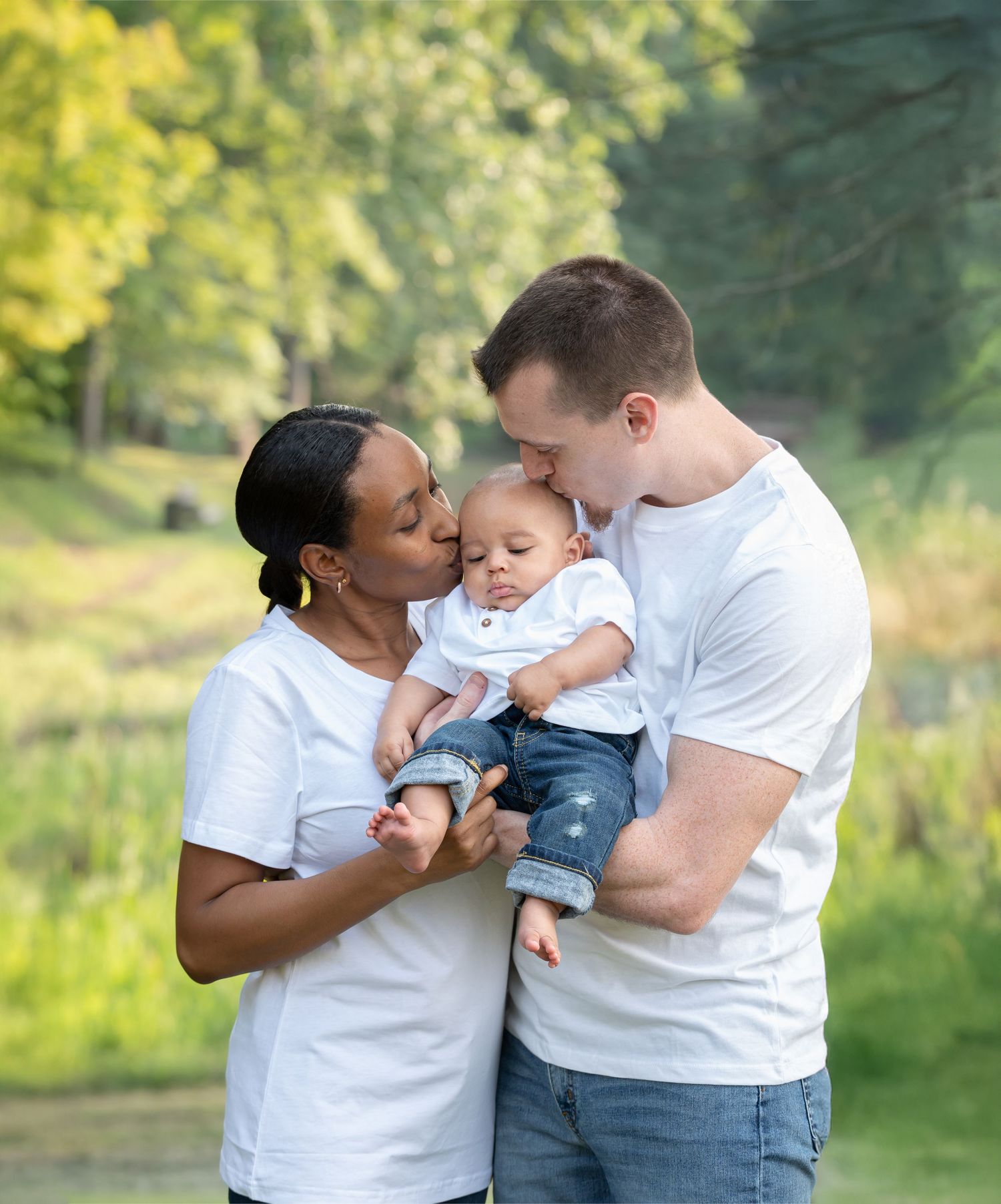 a young couple in white shirts holds their baby boy and kisses him on the head; soft green trees in the background, shot in Morgantown, WV