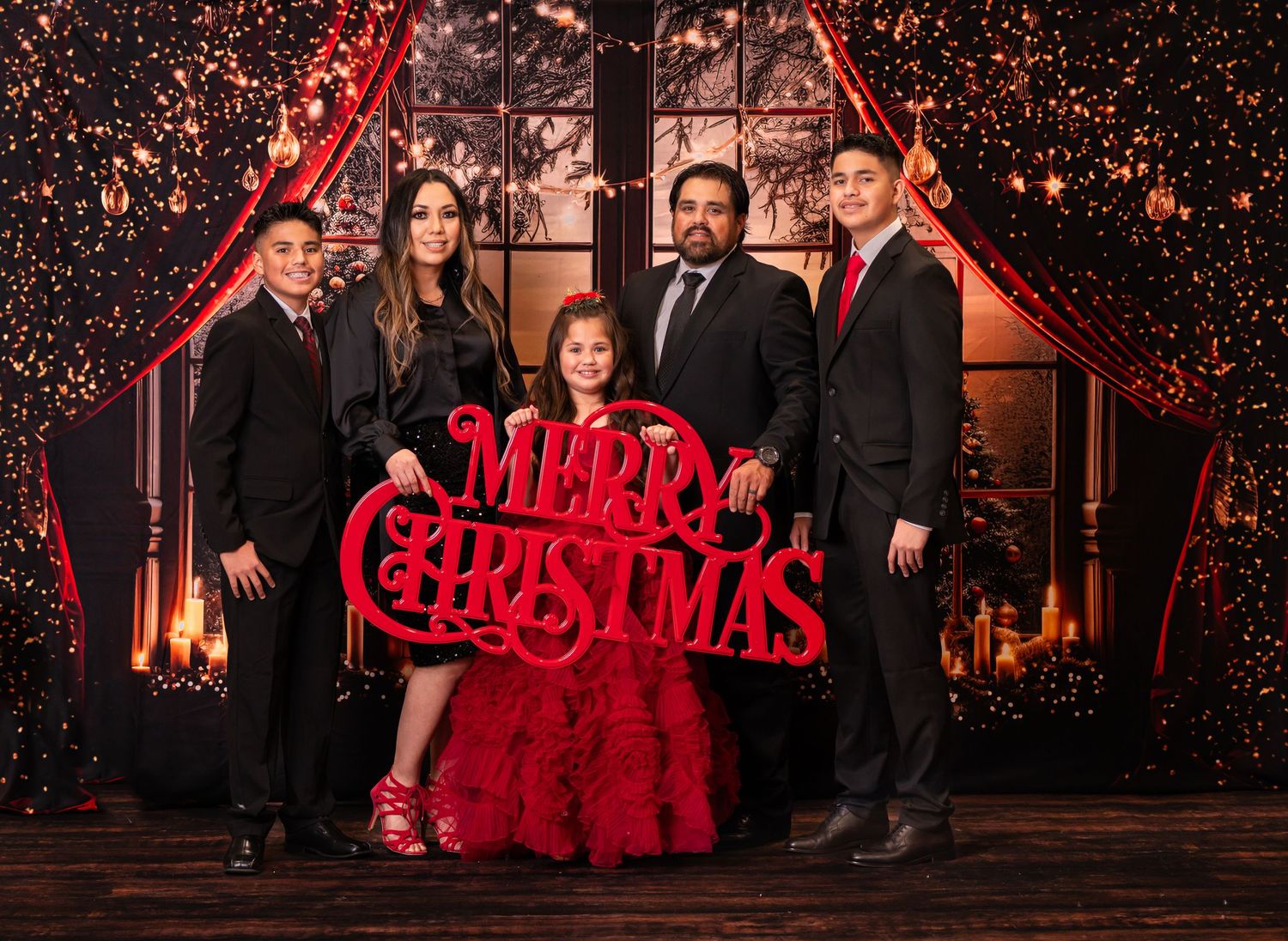 a family dressed in formal suits and dresses stand in front of deep red curtains lit with Christmas lights and candles & pose for a portrait