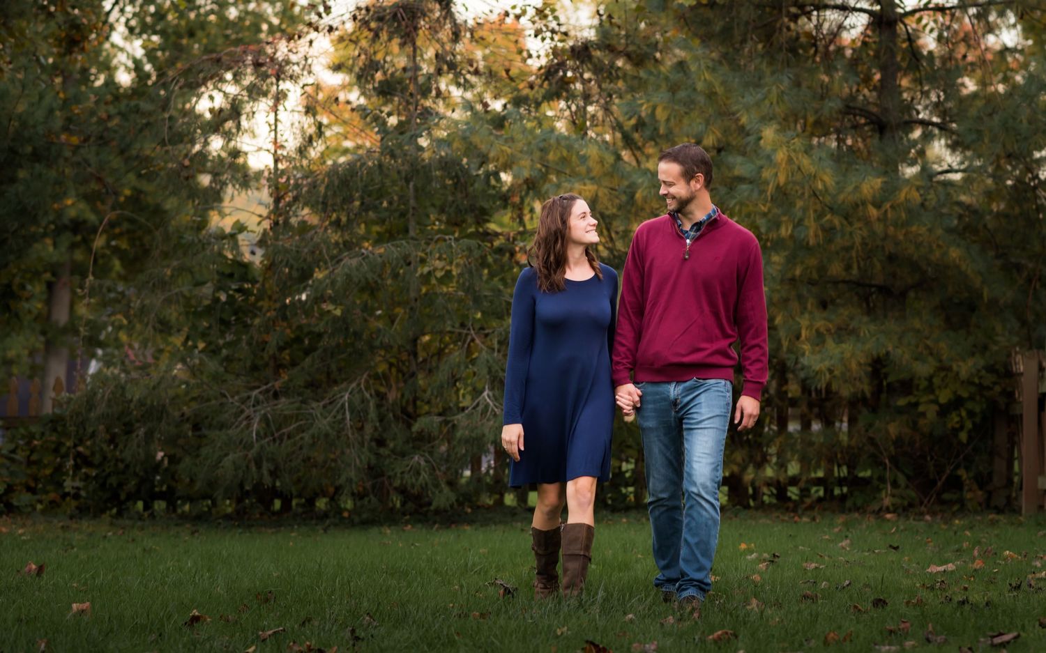 a husband and wife in winter sweaters stroll through the grass holding hands, and smile at one another