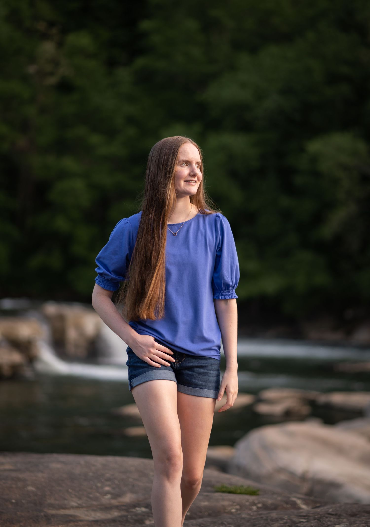 a high school graduate with long brown hair and a blue top walks toward the camera with a waterfall and large rocks behind her.
