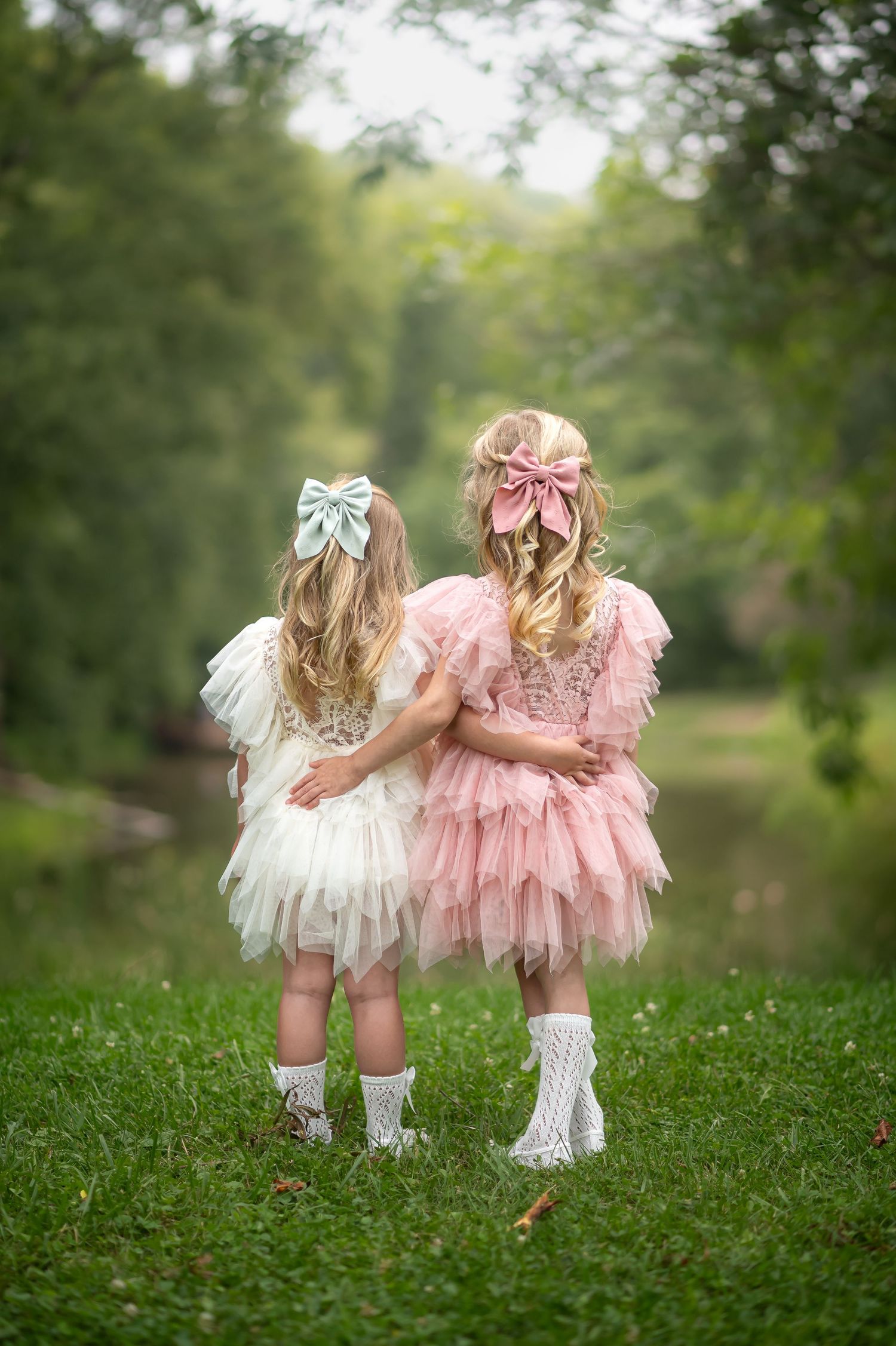 commercial product photo showing two little blonde girls in fancy pink and white dresses and bows, standing in a lush green meadow