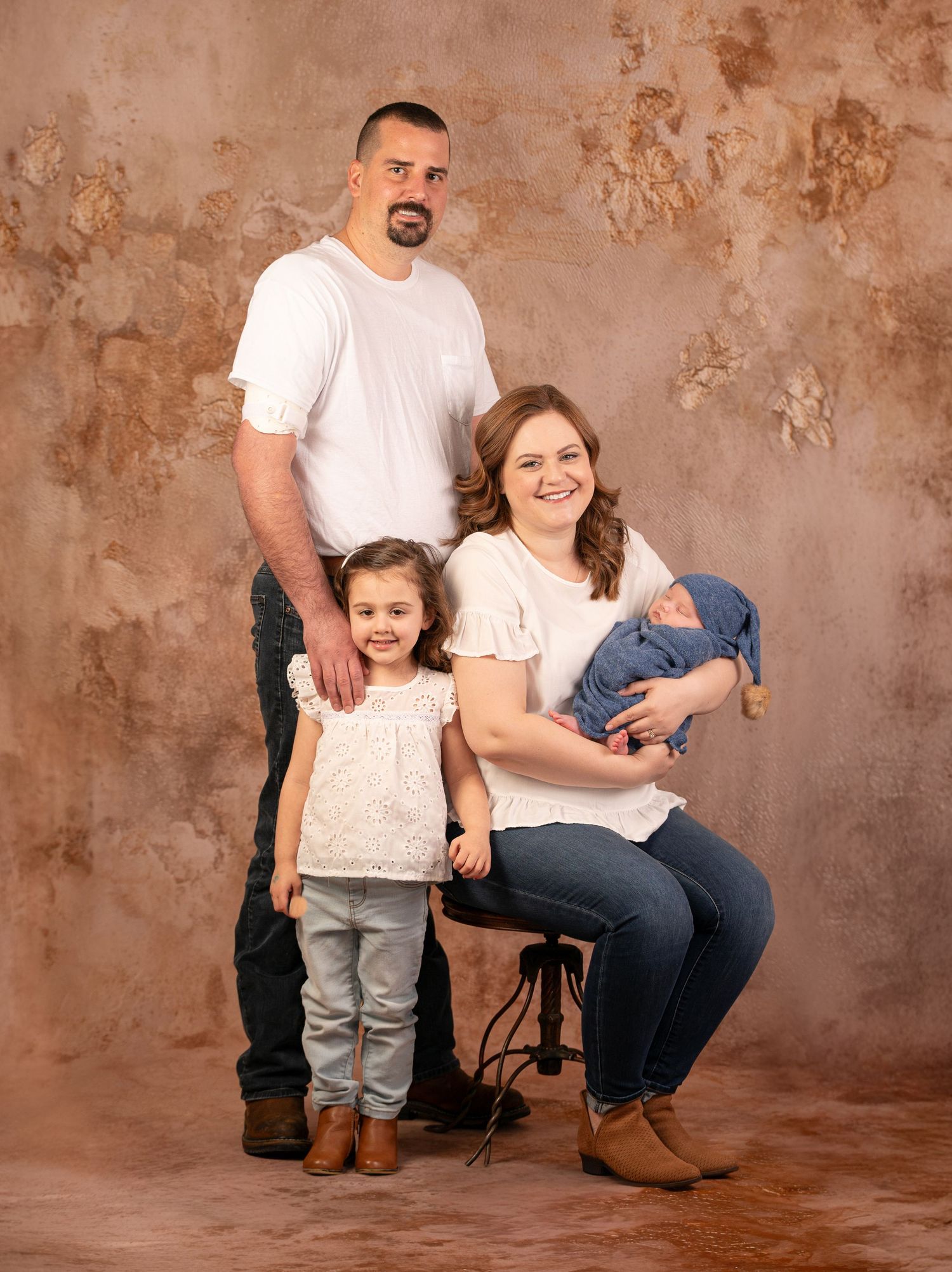 a family of four in white tshirts and jeans poses together for a portrait in a studio setting in front of a luxe brown and gold plaster drop
