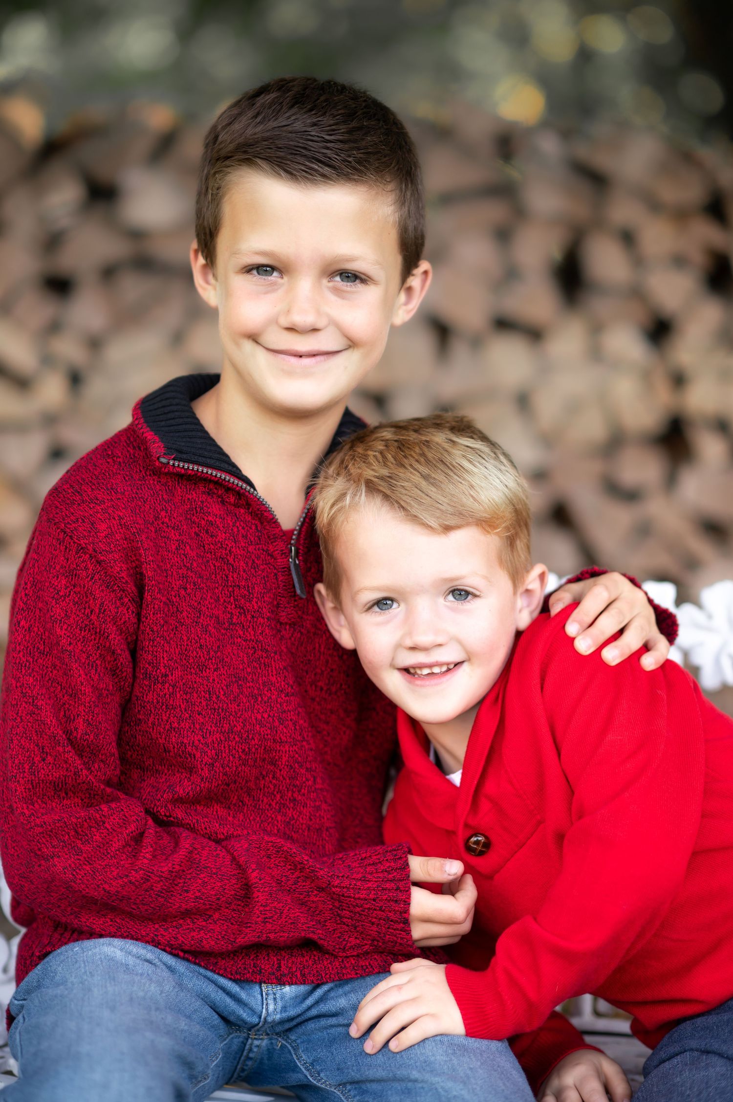 two little brothers in red sweaters pose for a Christmas portrait in front of stacked logs