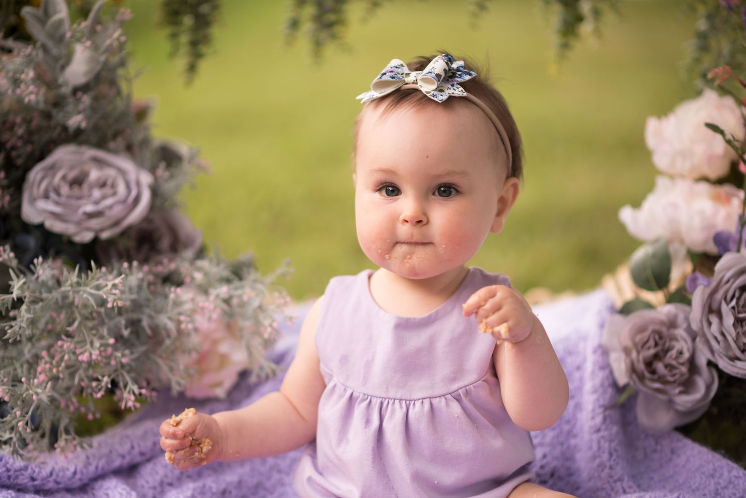 a chubby cheeked baby girl in a purple dress sits with her smash cake under an arch of purple and white flowers