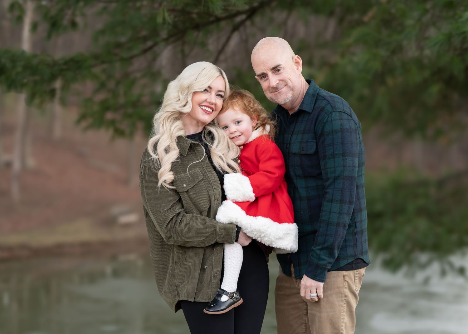 a father, mother and their little daughter pose for a Christmas card picture, surrounded by evergreen trees with a lake in the background