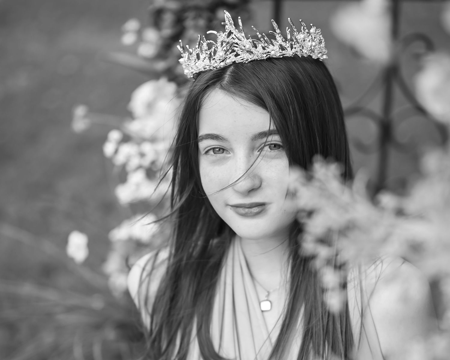 black and white closeup portrait of a pretty young girl with dark hair and freckles in a long dress and fairy crown, with flowers all around